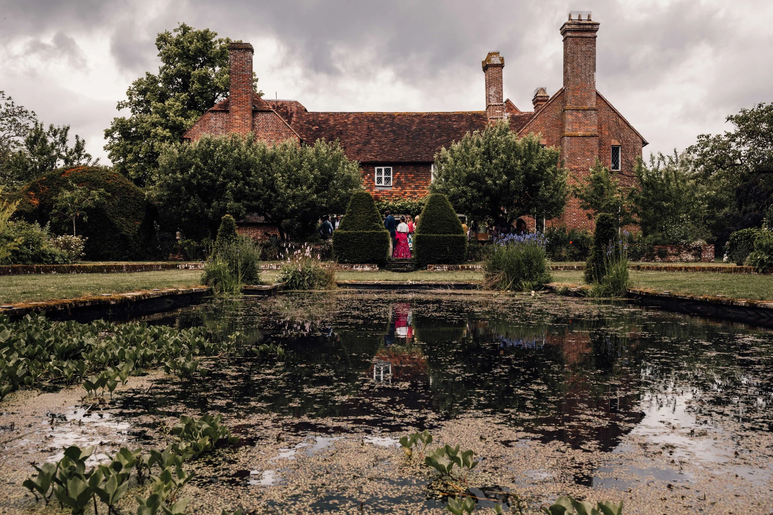 A historic brick house with a red tiled roof, multiple chimneys, and a large garden pond in the foreground, surrounded by trees and manicured bushes on a cloudy day.