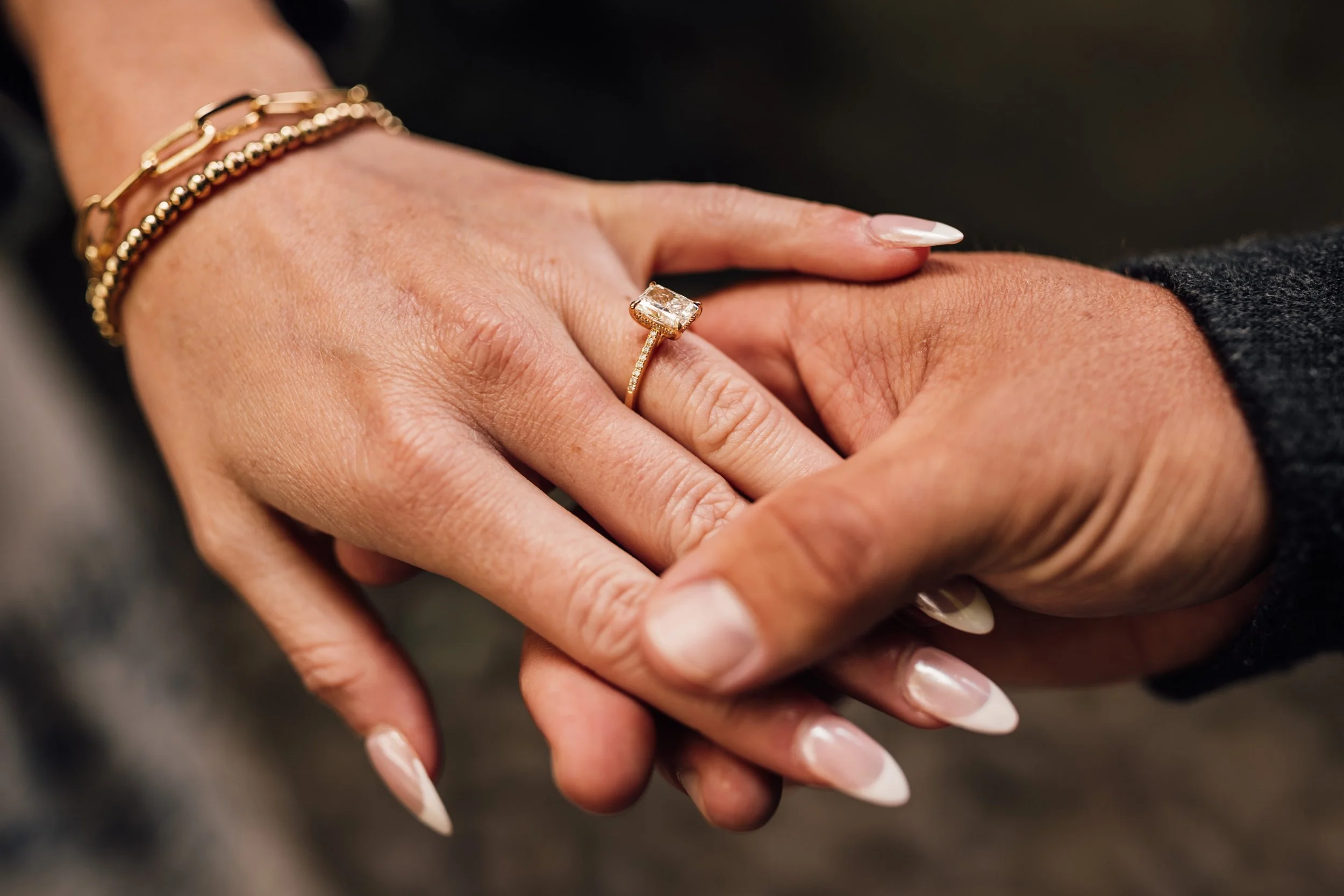 Close-up of a woman wearing a gold ring with a large gem, gently holding a man's hand. She has gold bracelets on her wrist and manicured nails.