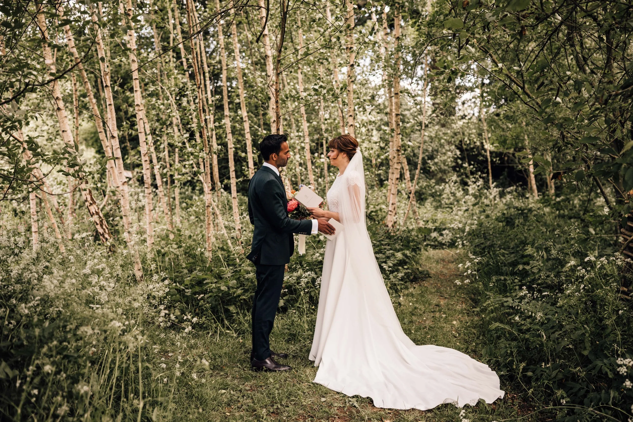A couple having a wedding ceremony in a wooded outdoor setting. The bride in a white gown is holding a bouquet and facing the groom dressed in a dark suit. They are standing close to each other, exchanging vows or rings, surrounded by green trees and
