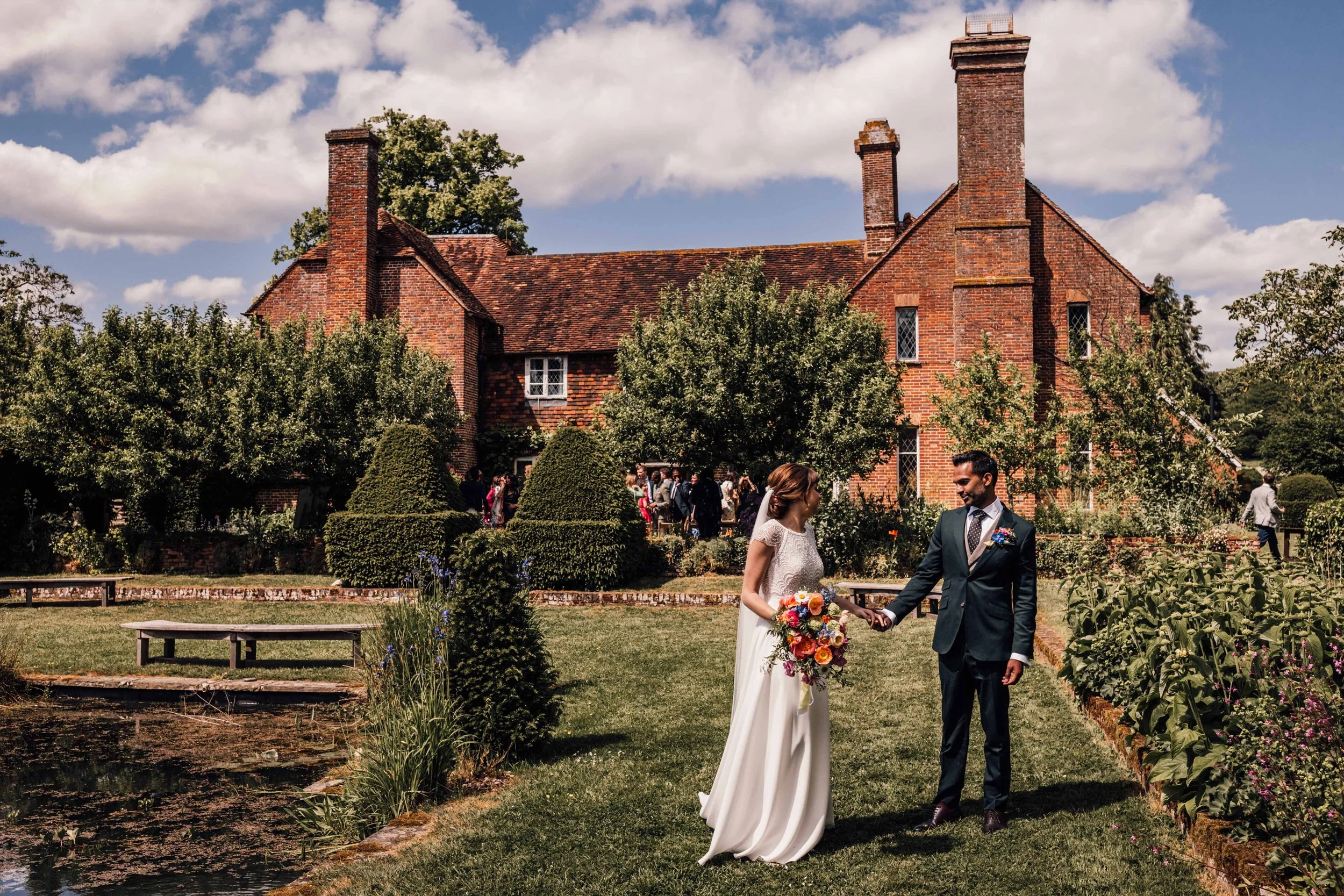 A bride and groom holding hands outside in a garden during a wedding ceremony. The bride is dressed in a white gown and holding a colorful bouquet, while the groom is dressed in a dark suit with a boutonniere. There is a large brick house in the back