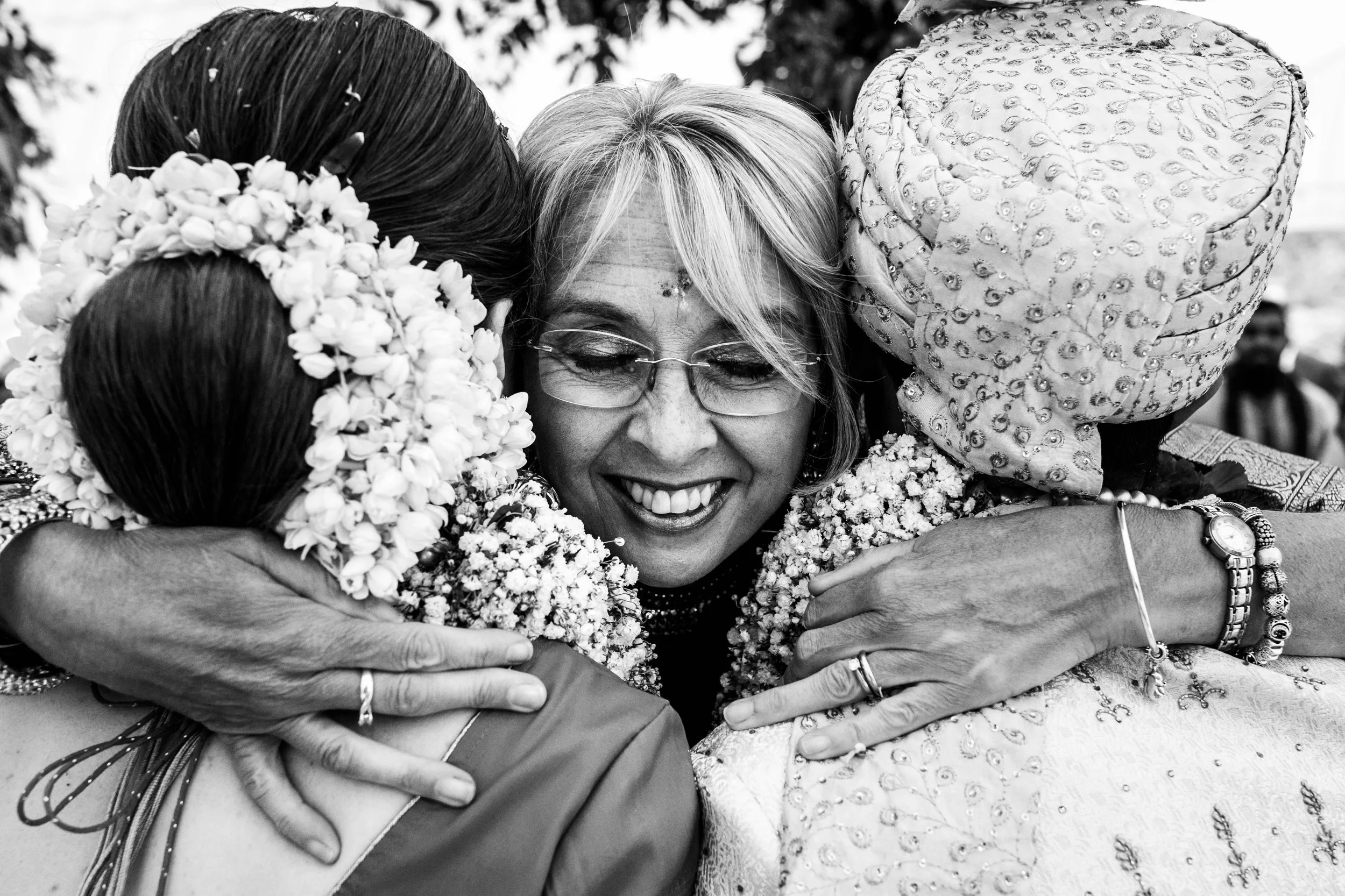 Three women embracing in hugs, smiling. One woman with glasses and gray hair is in the center, flanked by women wearing traditional head coverings and floral garlands.