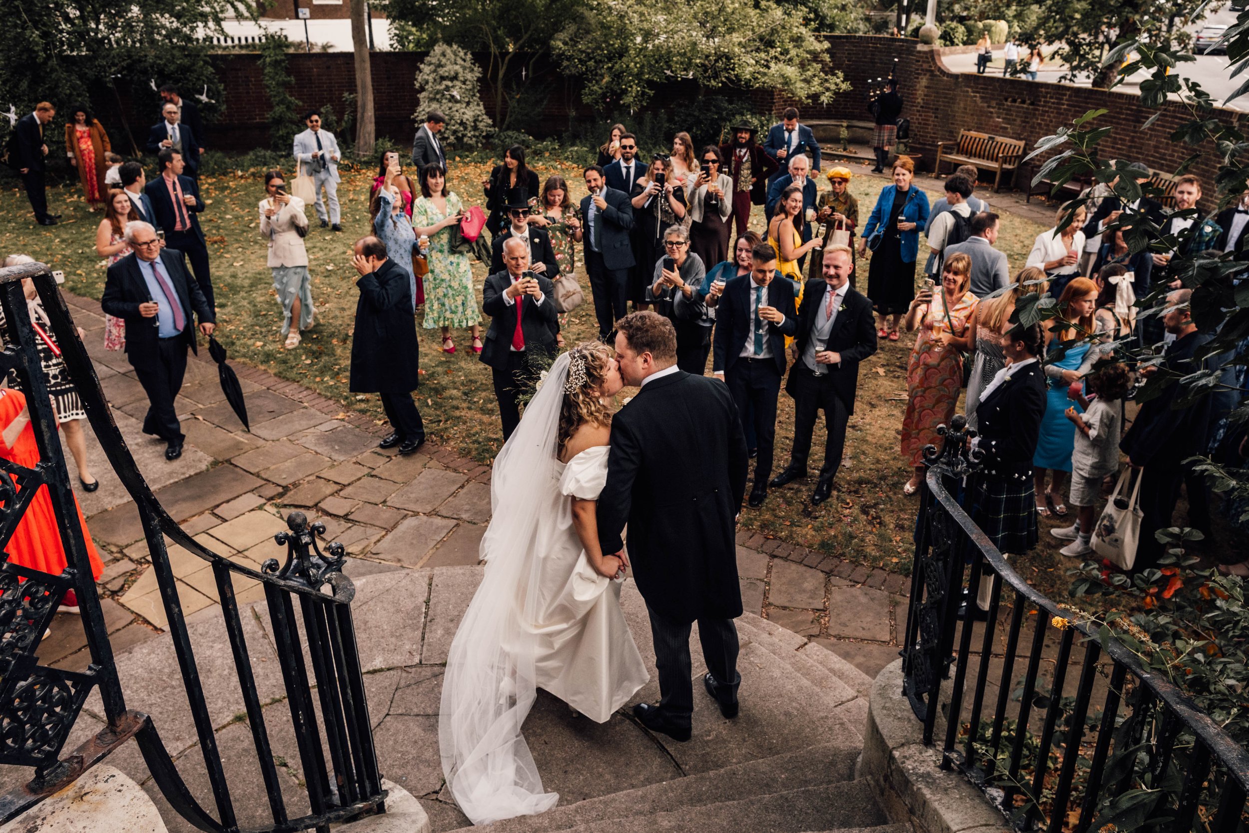 A bride and groom kissing on stone steps outside a wedding venue, surrounded by wedding guests taking photos and celebrating.