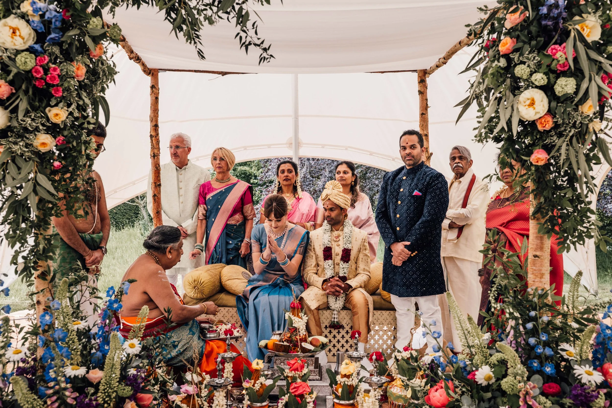 Indian multicultural wedding ceremony with bride and groom seated under floral canopy, surrounded by family and friends dressed in traditional attire, outdoors in a lush green setting.