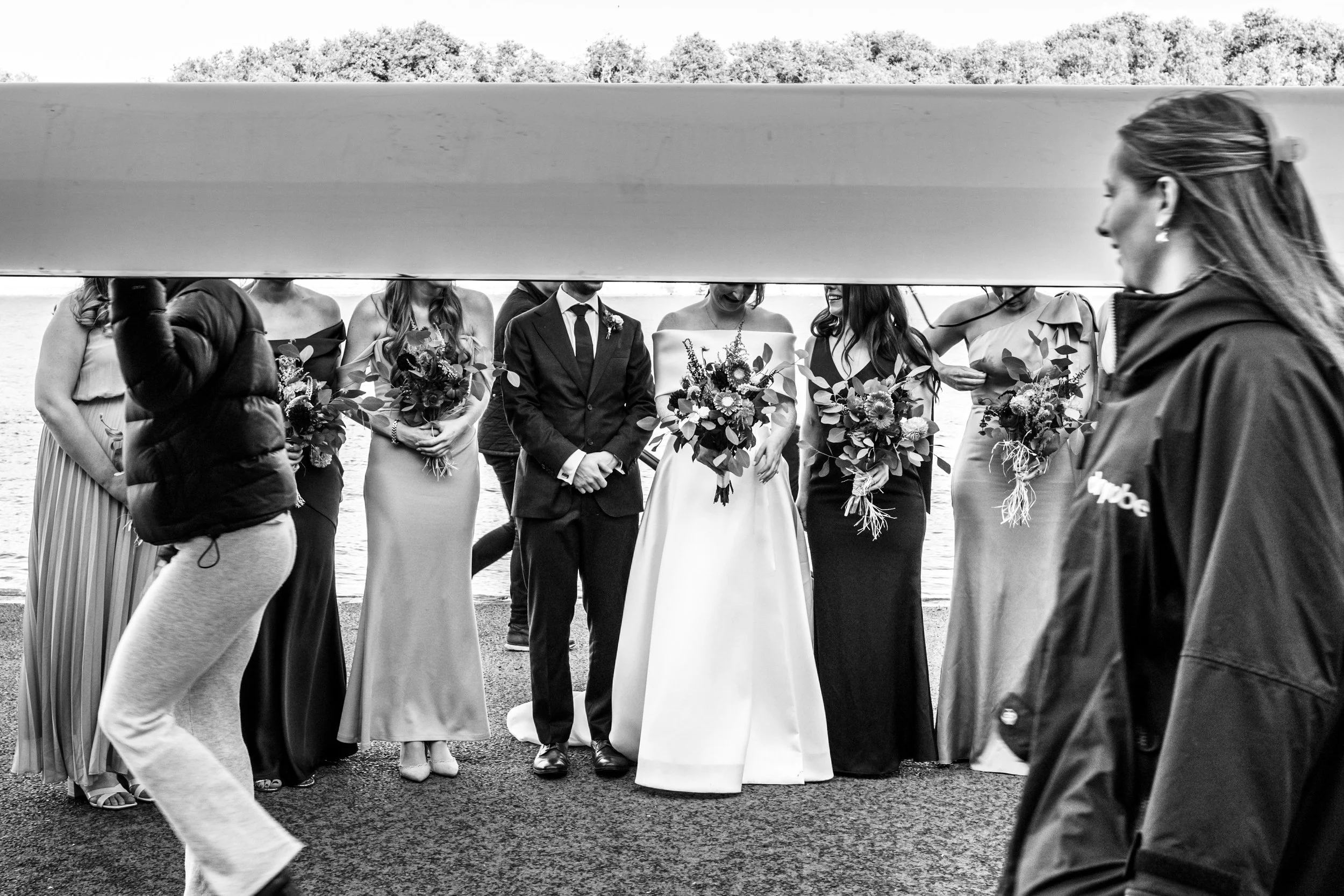 Black and white photo of a wedding party standing outdoors by a body of water, viewed from underneath a large horizontal object, with a woman on the right in a dark jacket and a puffy sleeve. The bride in a white gown and the groom in a dark suit are