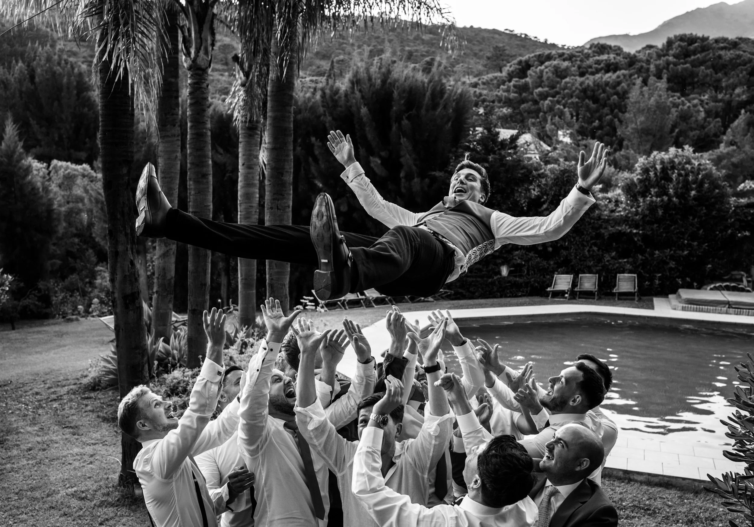 Group of men in business attire throwing a man into the air in an outdoor setting with trees and a swimming pool in the background