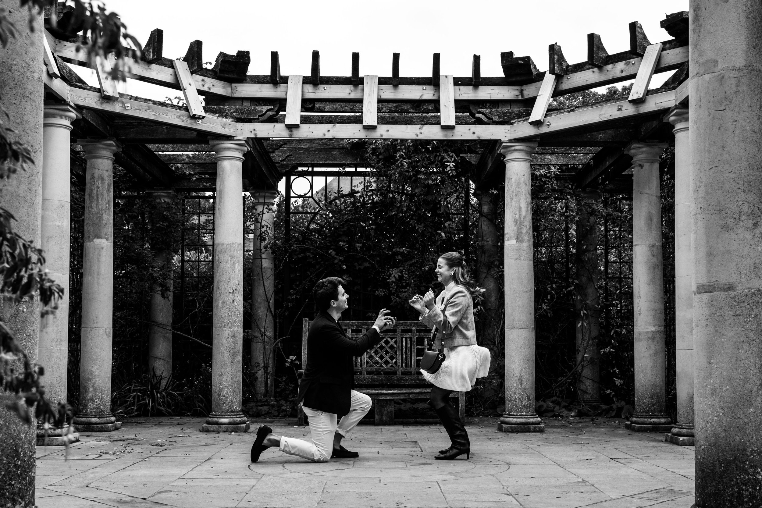 A man proposing to a woman in a garden with columns and a pergola, with the woman smiling and holding hands.