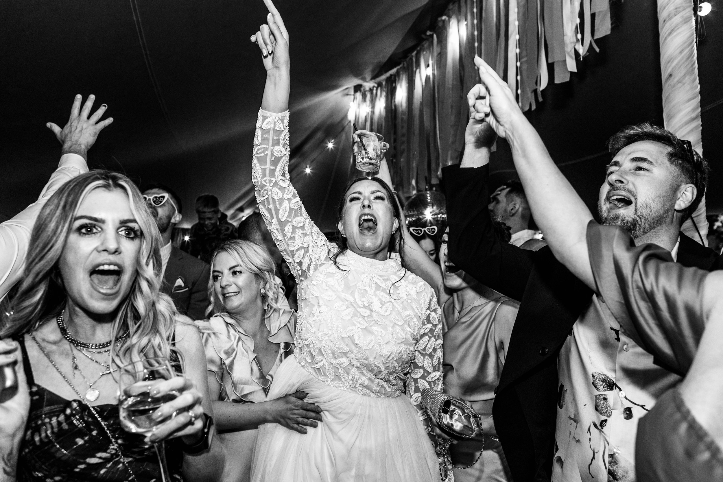 A group of people at a lively celebration or party, with a woman in a lace dress center, raising her arm, holding a drink, and shouting. Others around her are smiling, cheering, and dancing.
