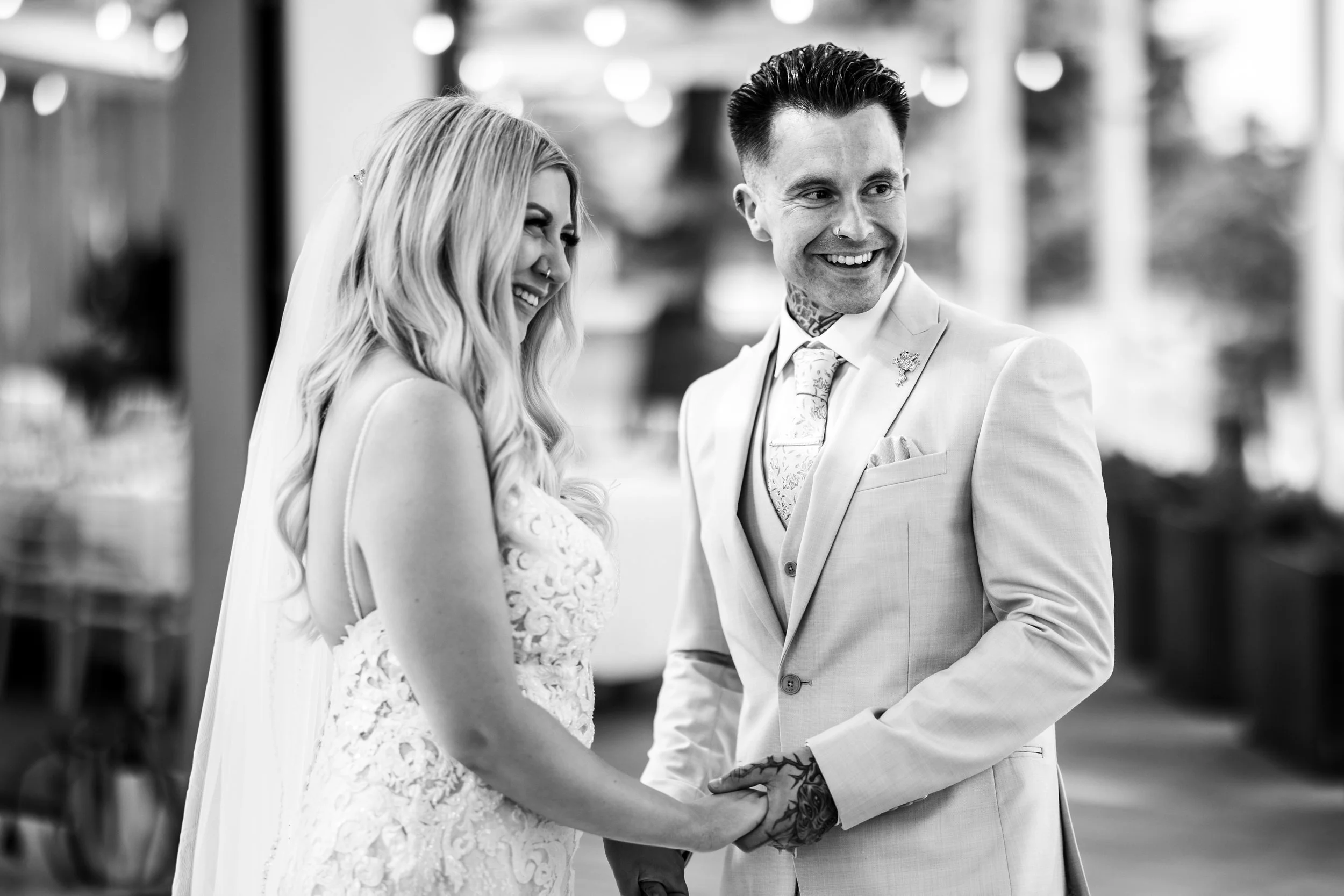 Black and white photo of a bride and groom holding hands, smiling, during their wedding ceremony. The bride is wearing a lace wedding gown and veil, and the groom is wearing a light-colored suit with a patterned tie. They are outdoors with a blurred 