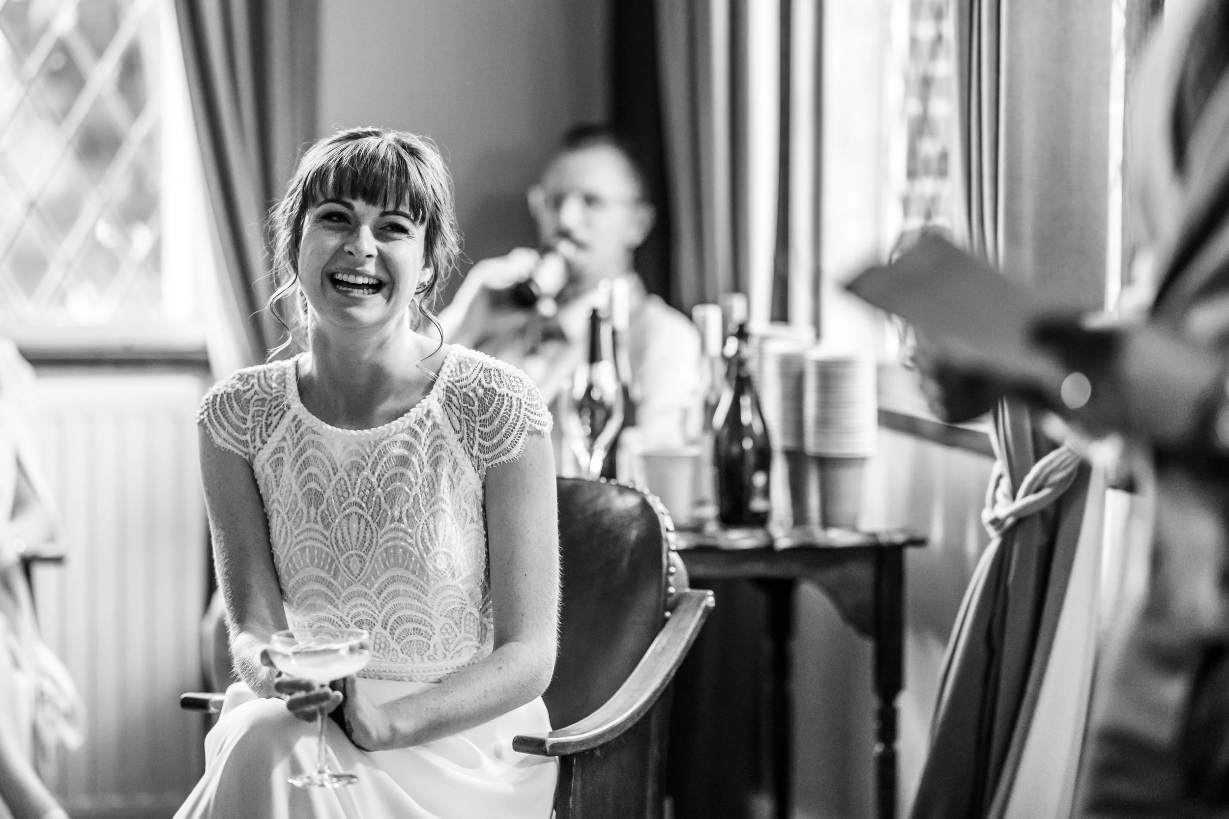 A woman with a big smile, holding a glass, sitting on a chair in a room with a window, with people and bottles in the background.