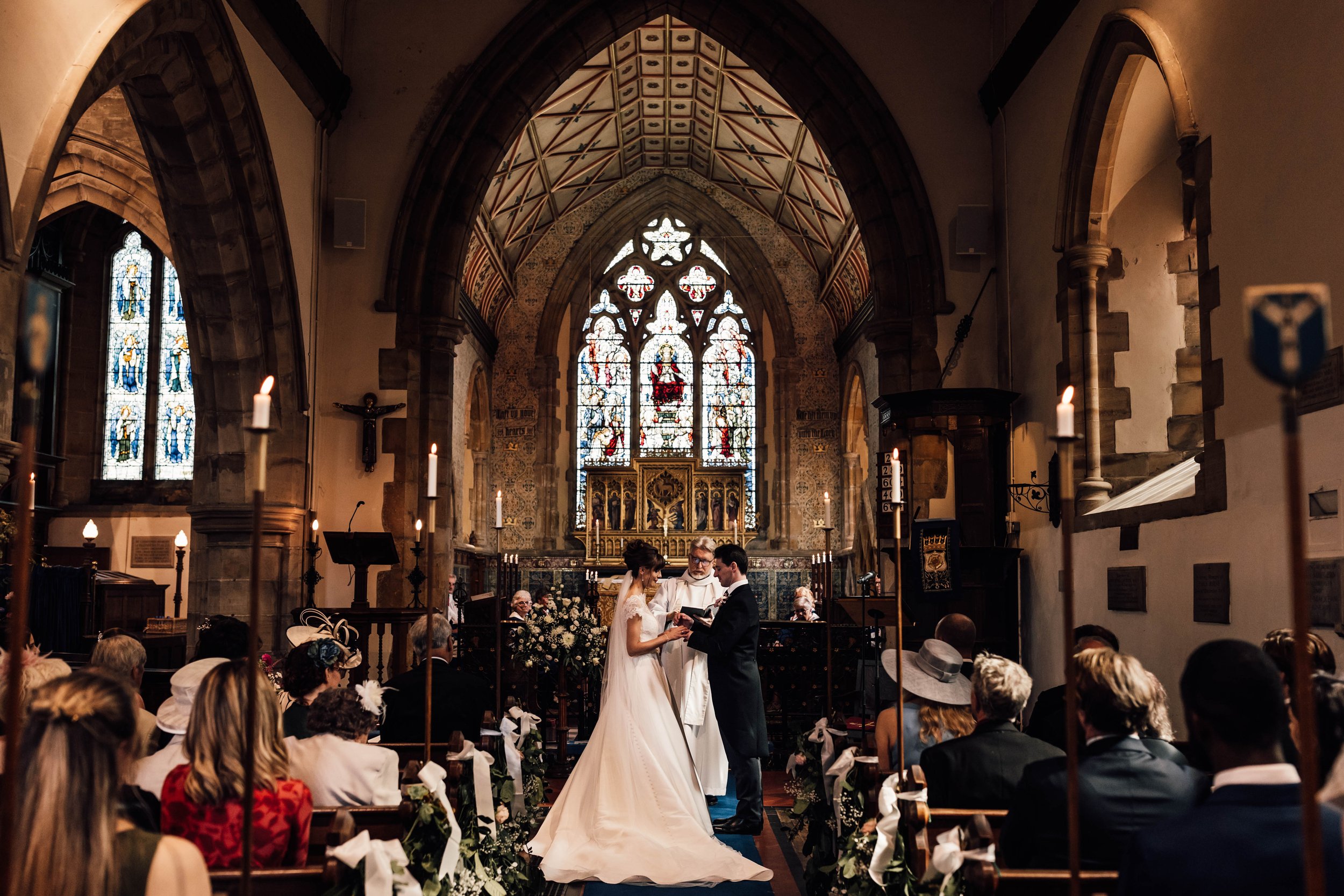 A wedding ceremony taking place inside a church with stained glass windows, a priest officiating, and the bride and groom exchanging vows in front of an altar, with guests seated on either side.