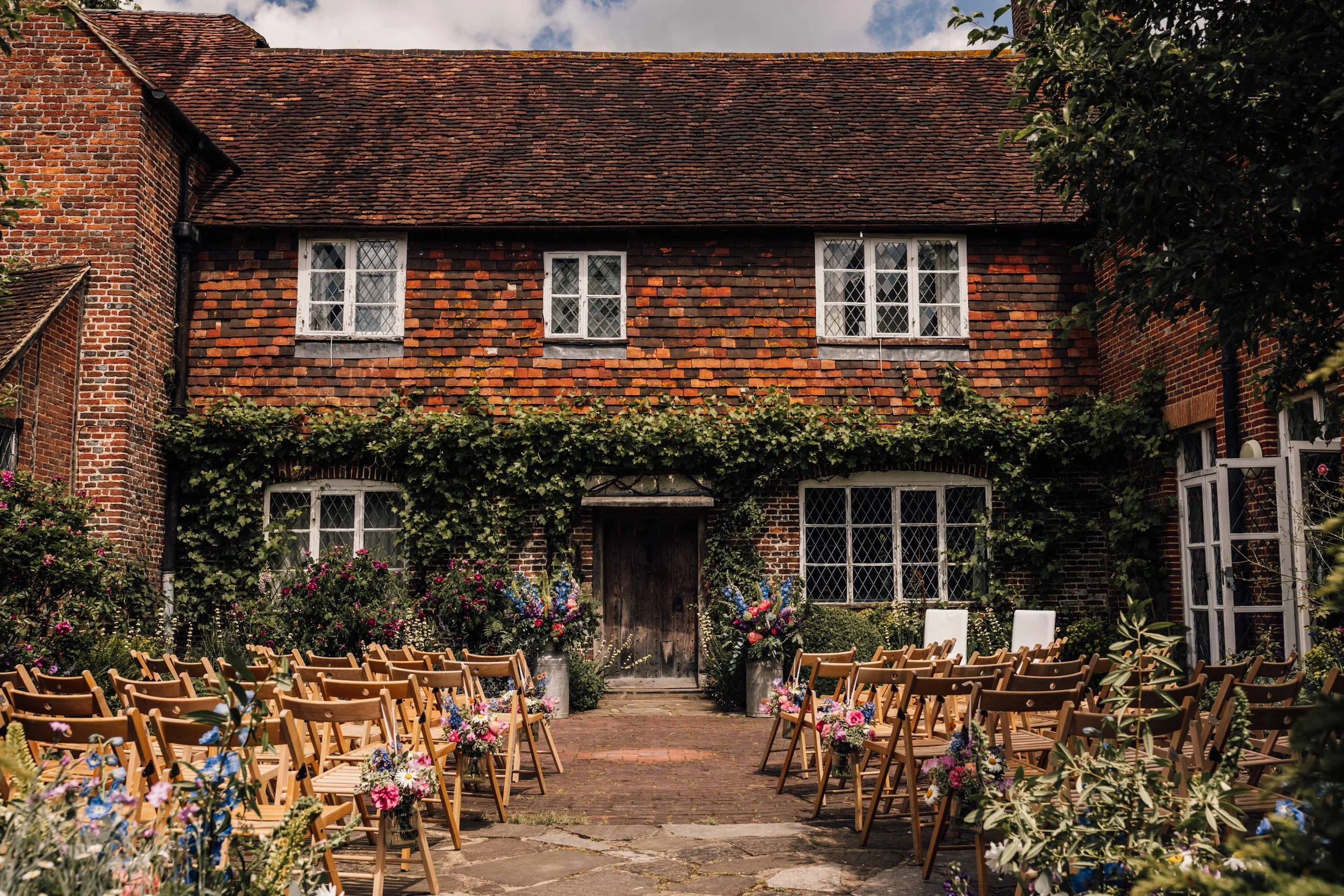 Outdoor wedding setup with rows of wooden chairs decorated with flowers, in front of a brick building with ivy and flowering plants.