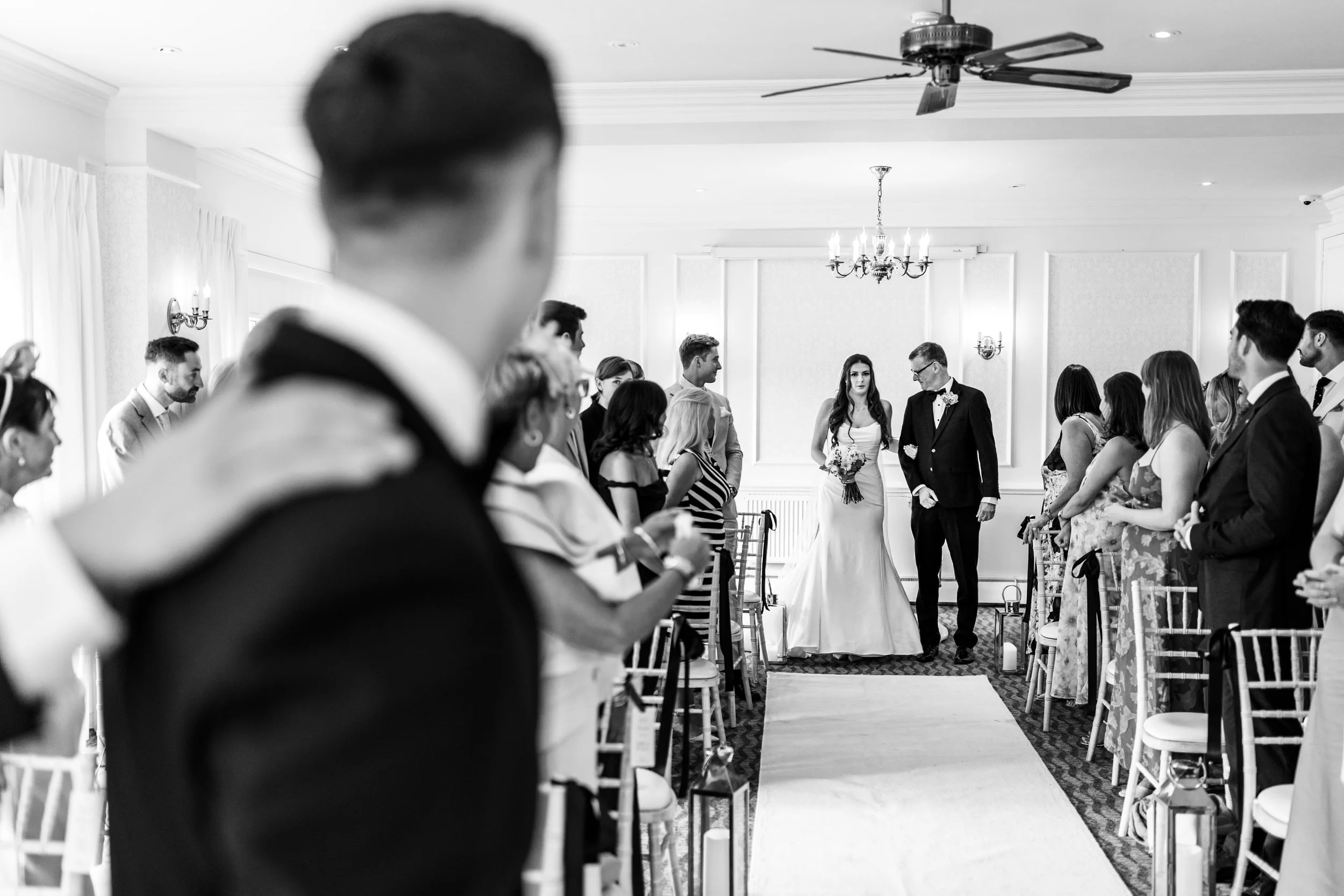 Black and white photo of a wedding ceremony with the bride walking down the aisle, accompanied by a man, likely her father, with guests seated on either side.