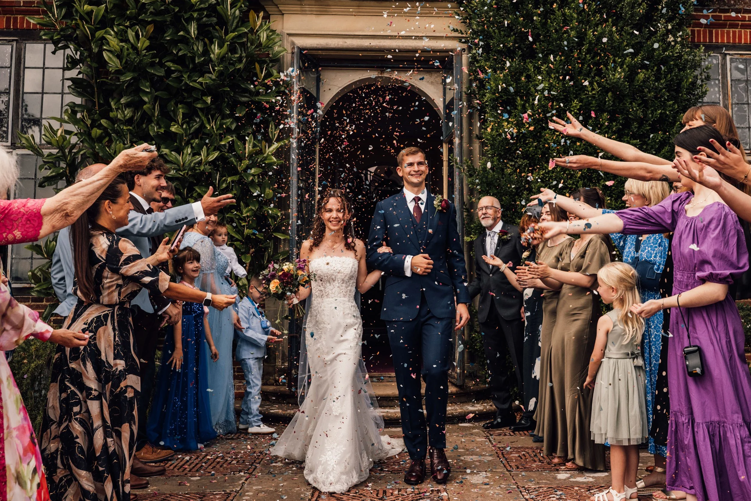 A newlywed couple walks arm-in-arm out of a church while guests throw confetti to celebrate their wedding.