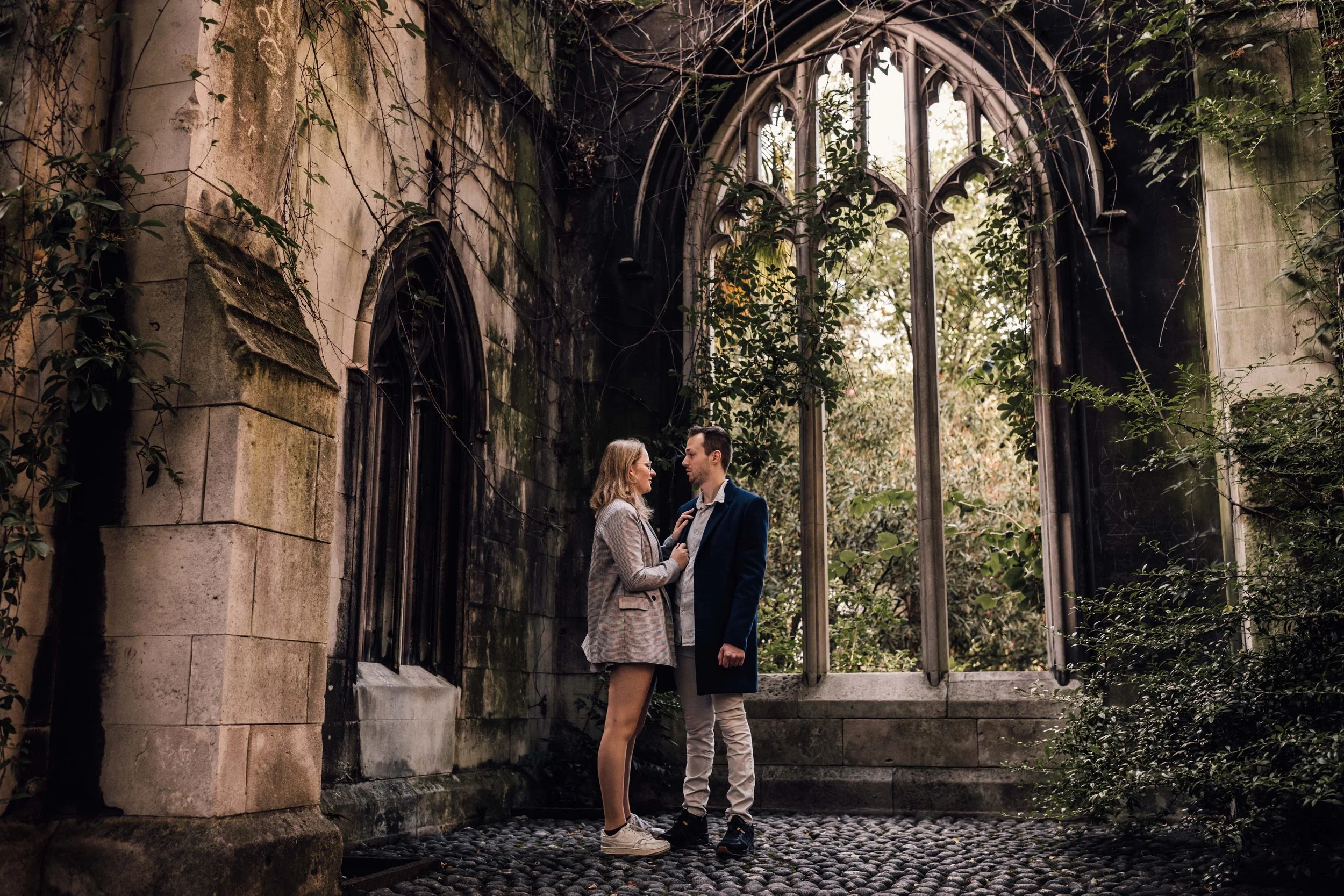 A couple standing close together in front of a large, old stained glass window in a ruined church or chapel, surrounded by greenery and vines.