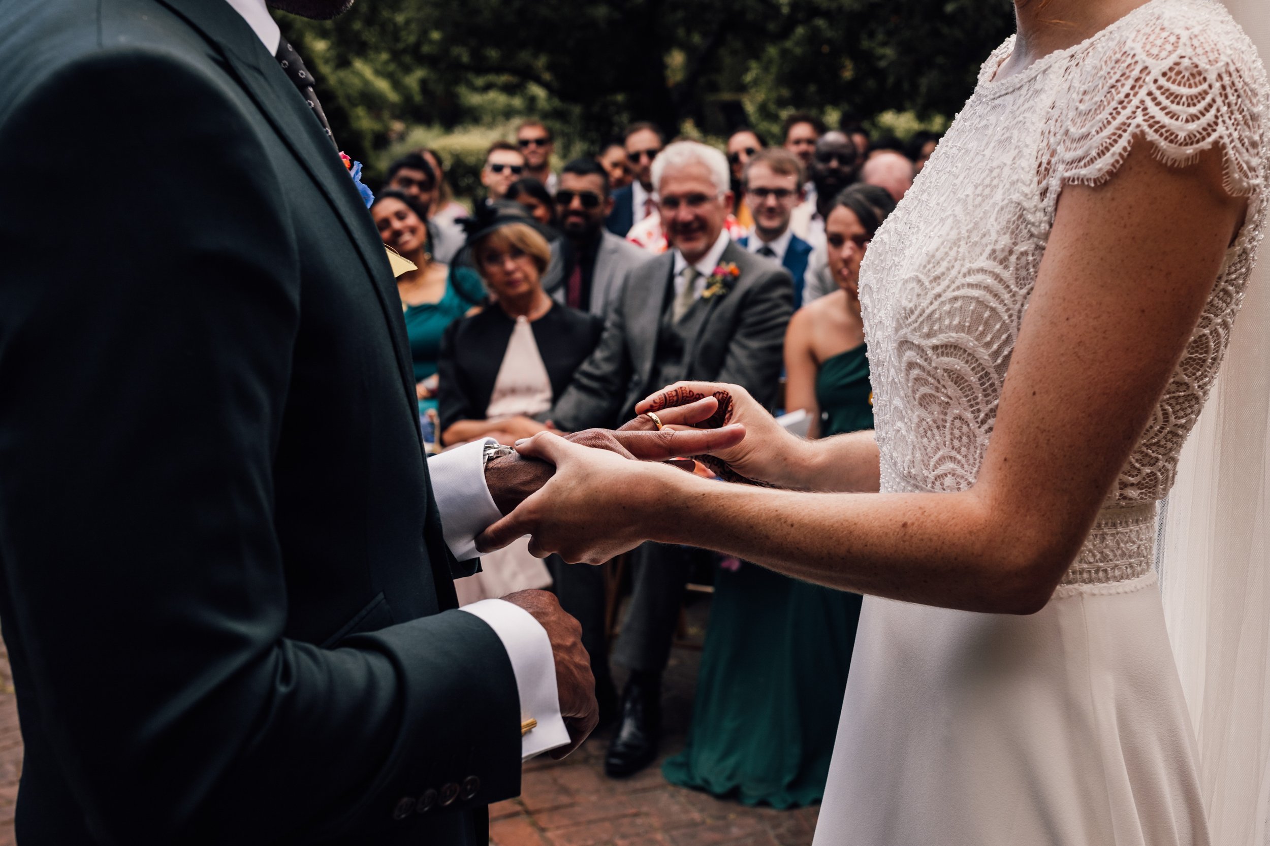 A bride and groom exchanging rings during an outdoor wedding ceremony, with guests watching in the background.