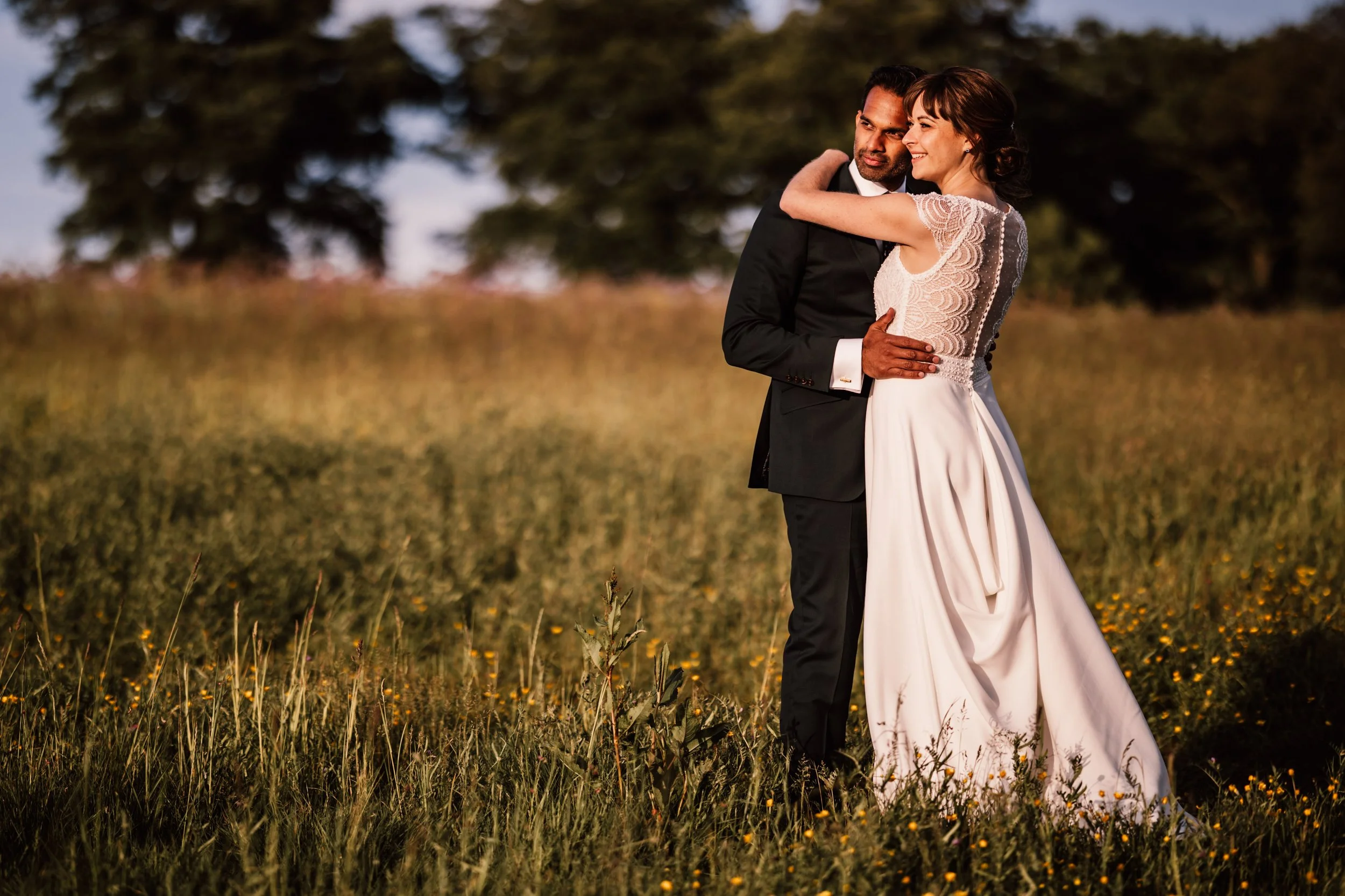 A couple dressed in wedding attire embracing in a field of wildflowers at sunset.
