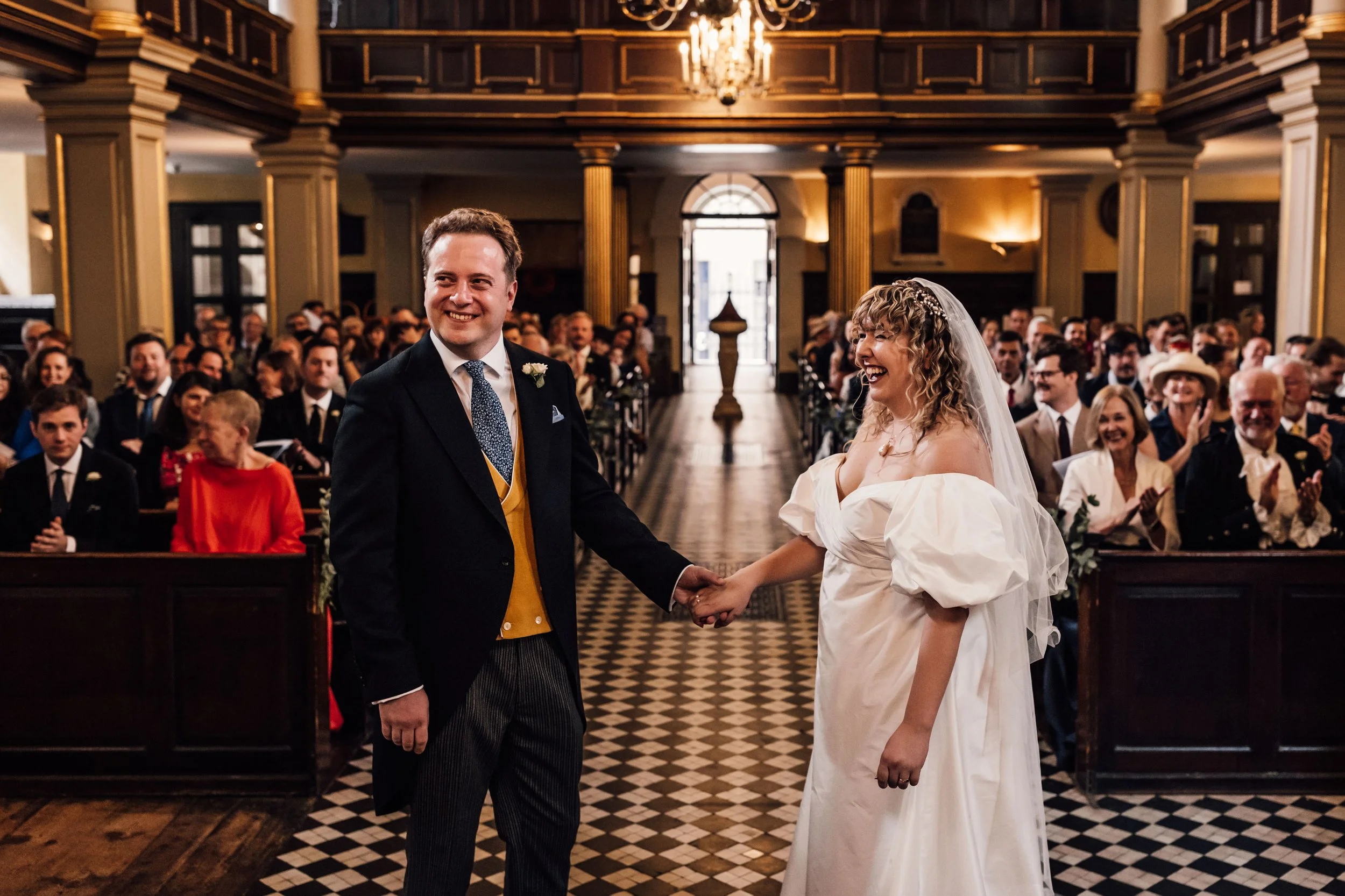 A couple exchanges vows at their wedding ceremony inside a decorated hall with guests watching and applauding.
