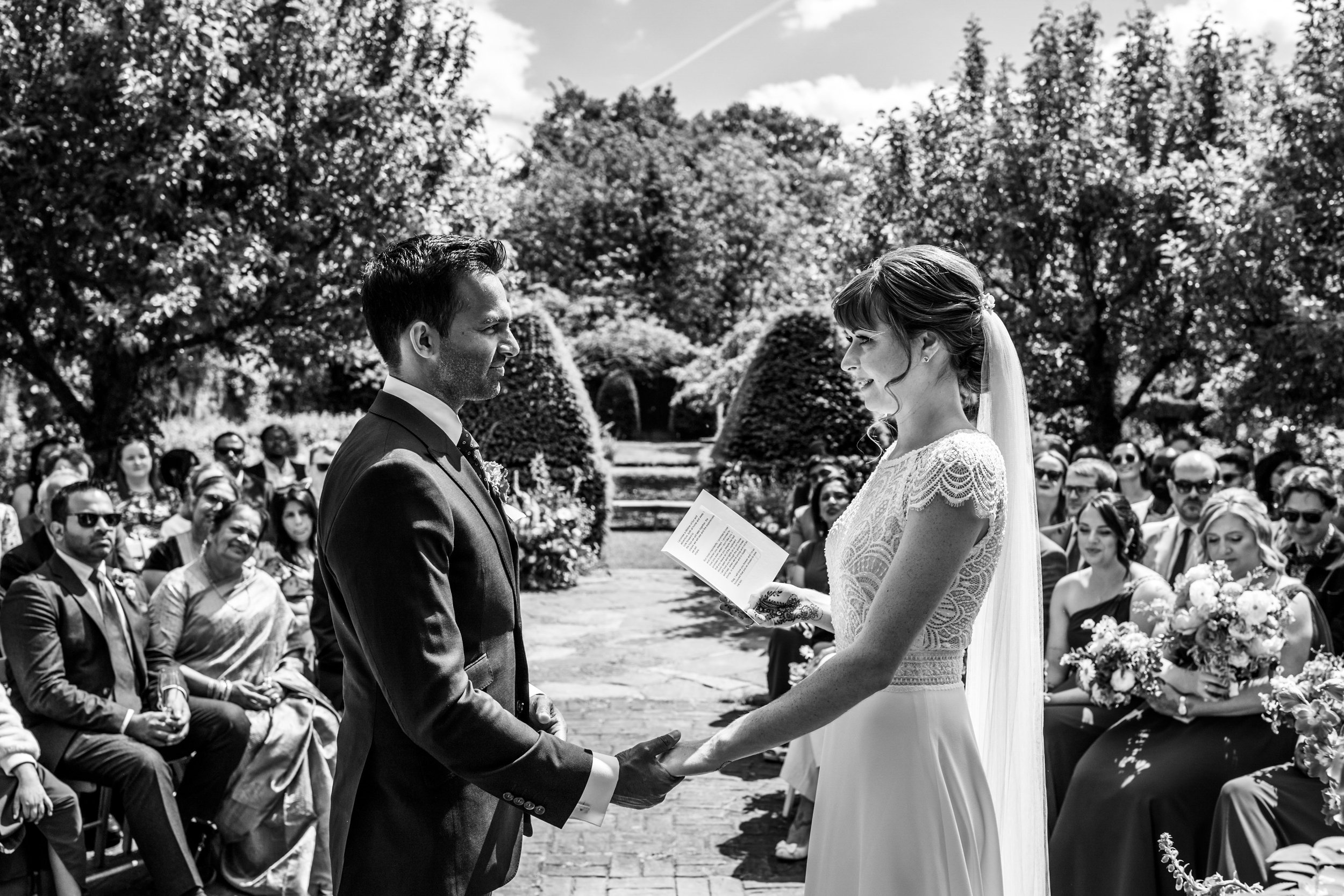 Black and white wedding ceremony outdoors with a bride and groom holding hands, facing each other, surrounded by seated guests under trees.