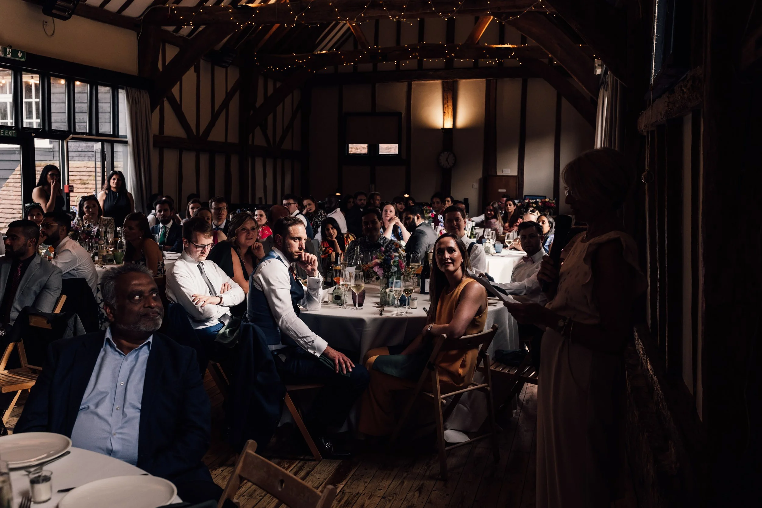 A woman in a cream-colored dress speaking into a microphone at a formal event held in a rustic venue, with many seated guests listening.