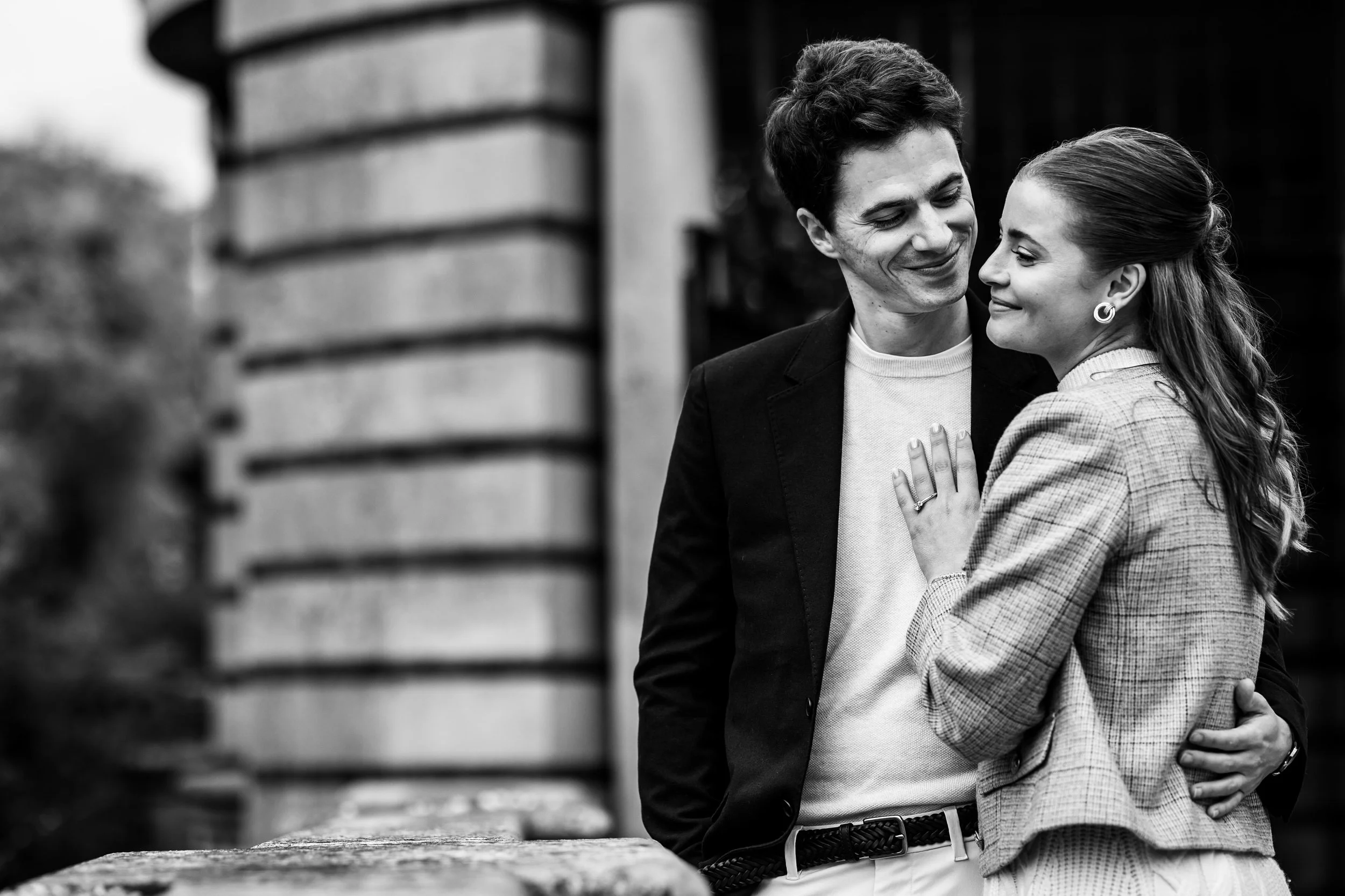 A black and white photo of a couple embracing outdoors, with the woman touching the man's chest and both smiling softly. The scene includes a blurred brick structure in the background.