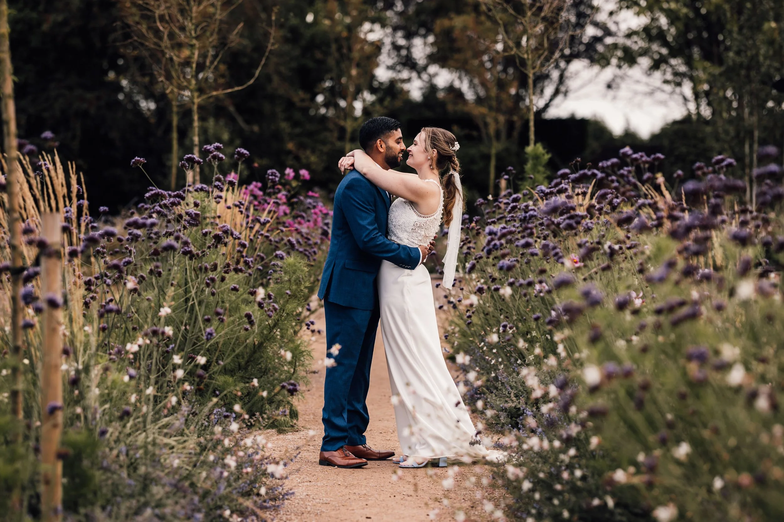 A bride and groom embrace on a dirt path surrounded by purple and pink flowers in a garden setting.