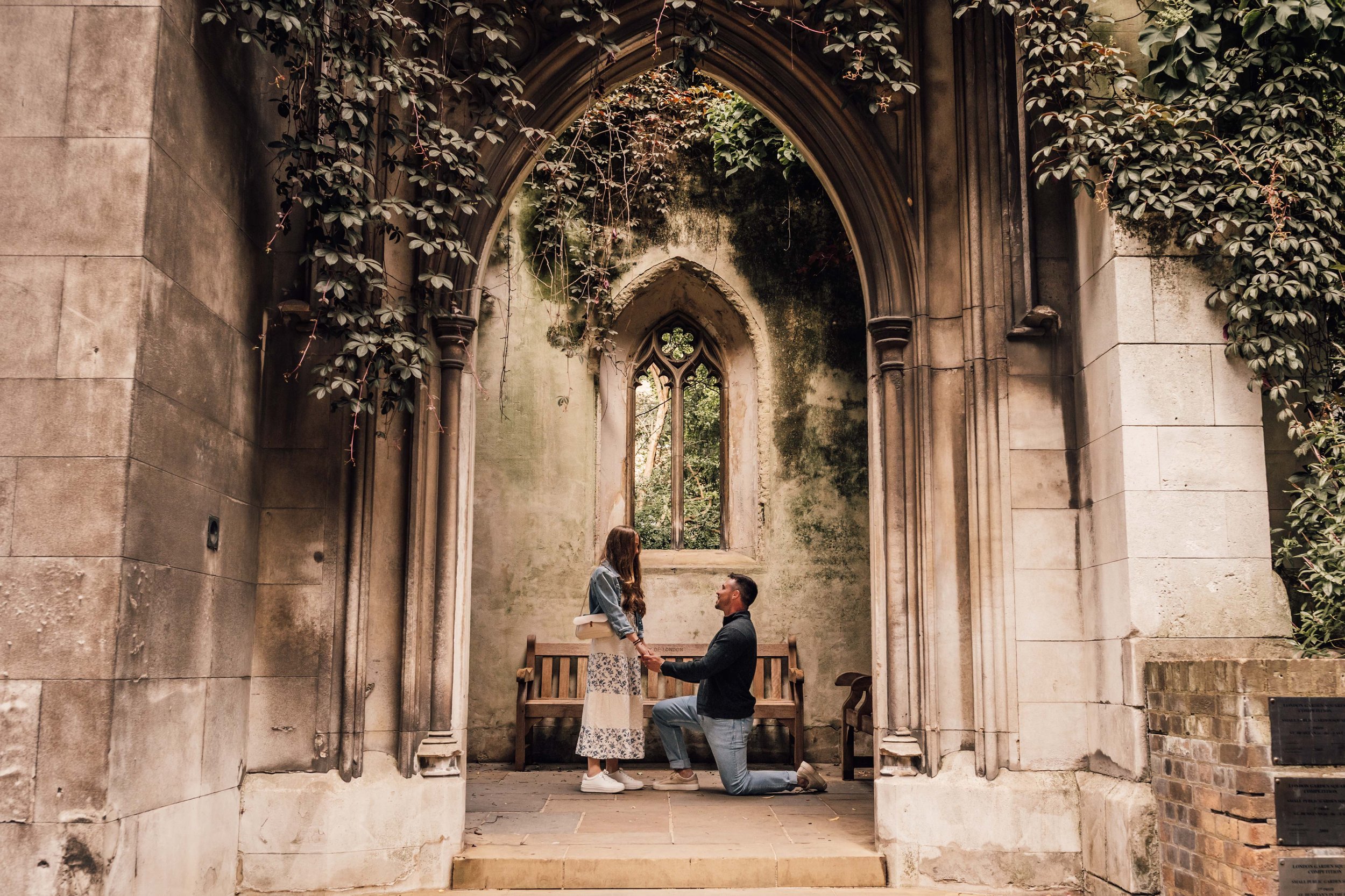A couple is in a romantic proposal scene inside an old, weathered stone church with an arched window and overgrown vines, where the man is kneeling and holding the woman’s hands.