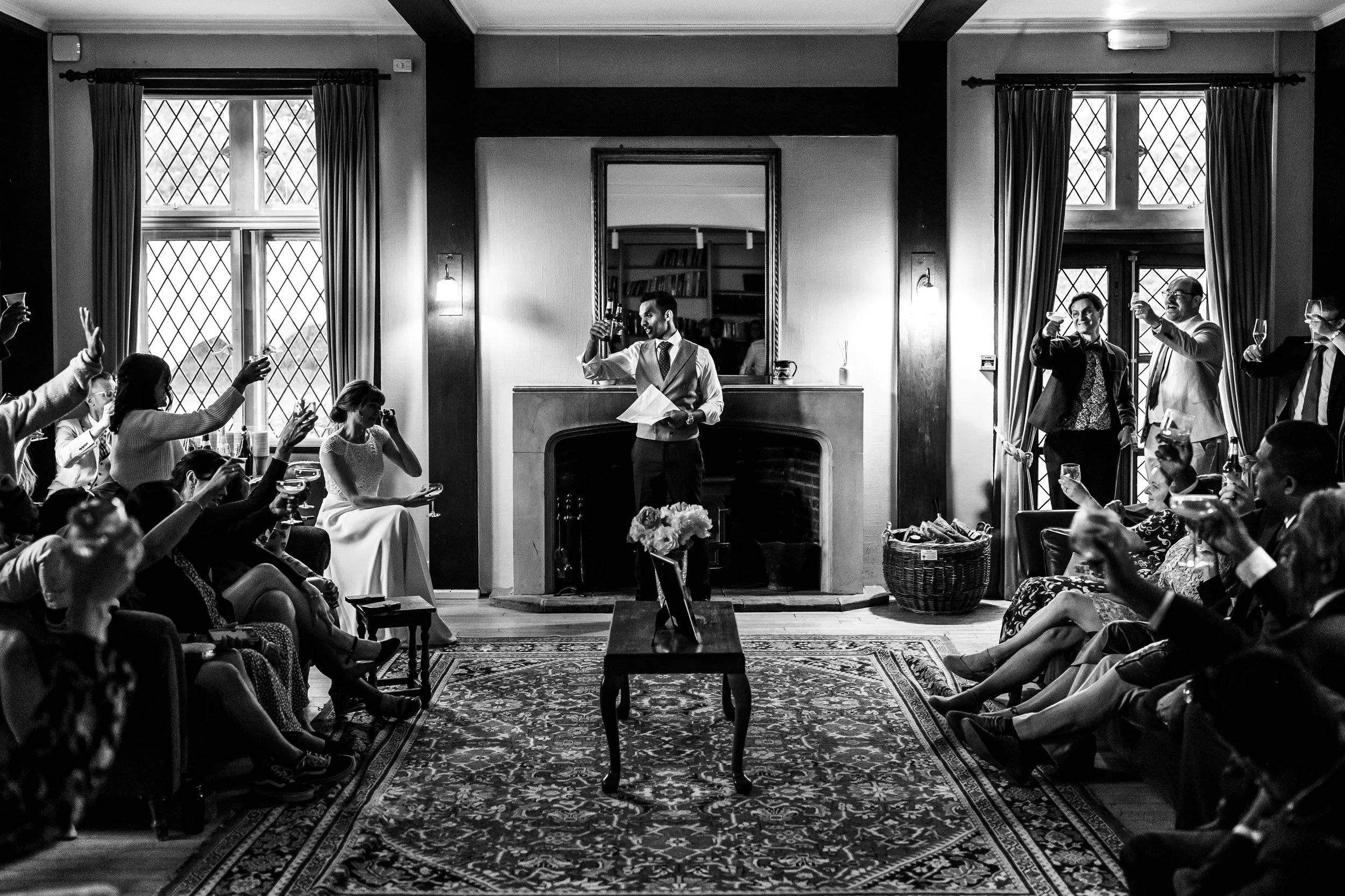 A black-and-white photo of a wedding toast in a cozy living room. The bride is seated on the left, covering her face with her hand, while the groom stands in the center raising a drink. Guests surround them, sitting and standing, raising glasses, and