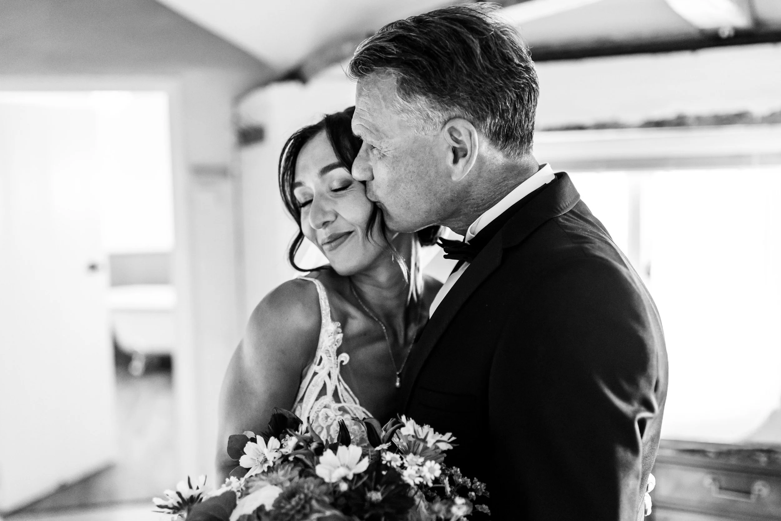 Black and white photo of a wedding couple, the man kissing the woman's forehead as she holds a bouquet, in an indoor setting with bright windows.