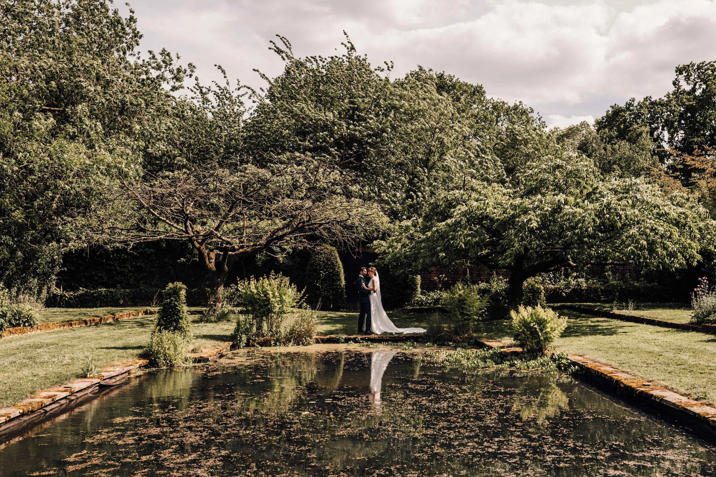 A bride and groom standing together in a garden, surrounded by trees and greenery, with a pond in the foreground and their reflection visible in the water.