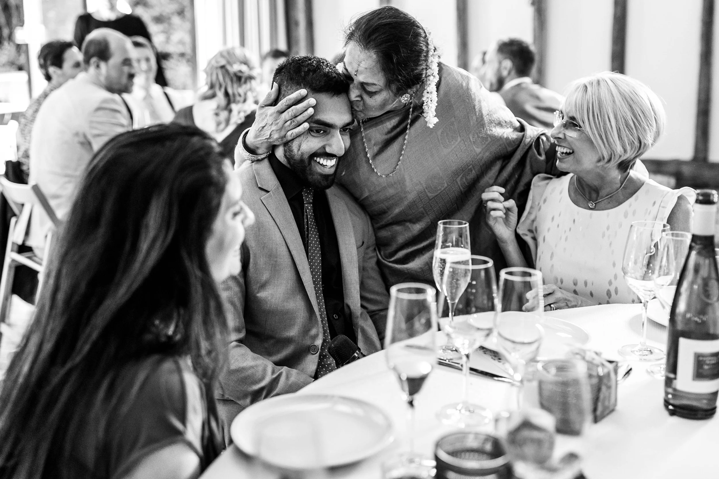 A group of people celebrating at a dining table, with an elderly woman kissing a smiling young man on the forehead, surrounded by glasses of champagne and bottles, in a joyful and warm atmosphere.