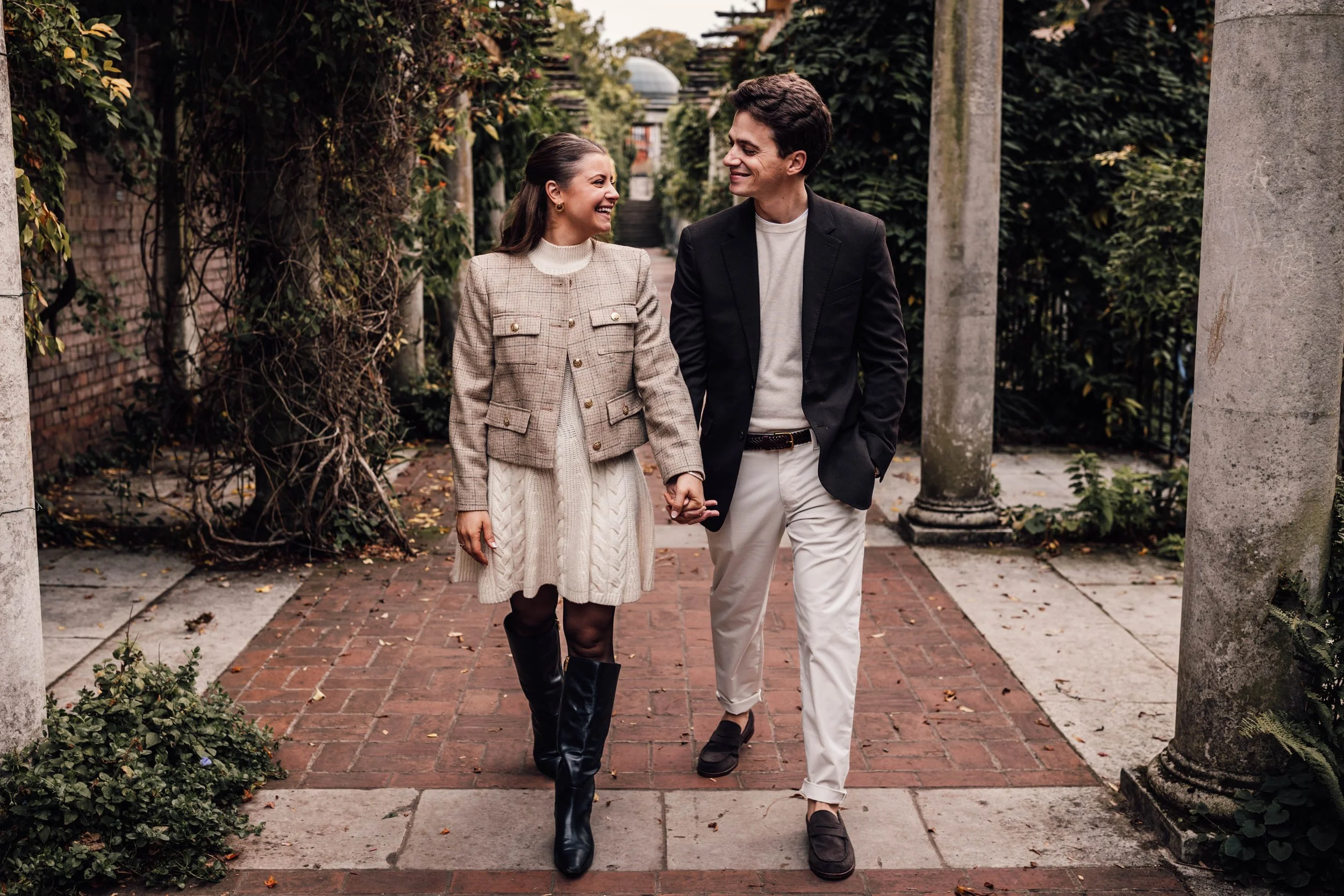 A smiling couple walks hand-in-hand outdoors on a brick path surrounded by greenery, columns, and trees
