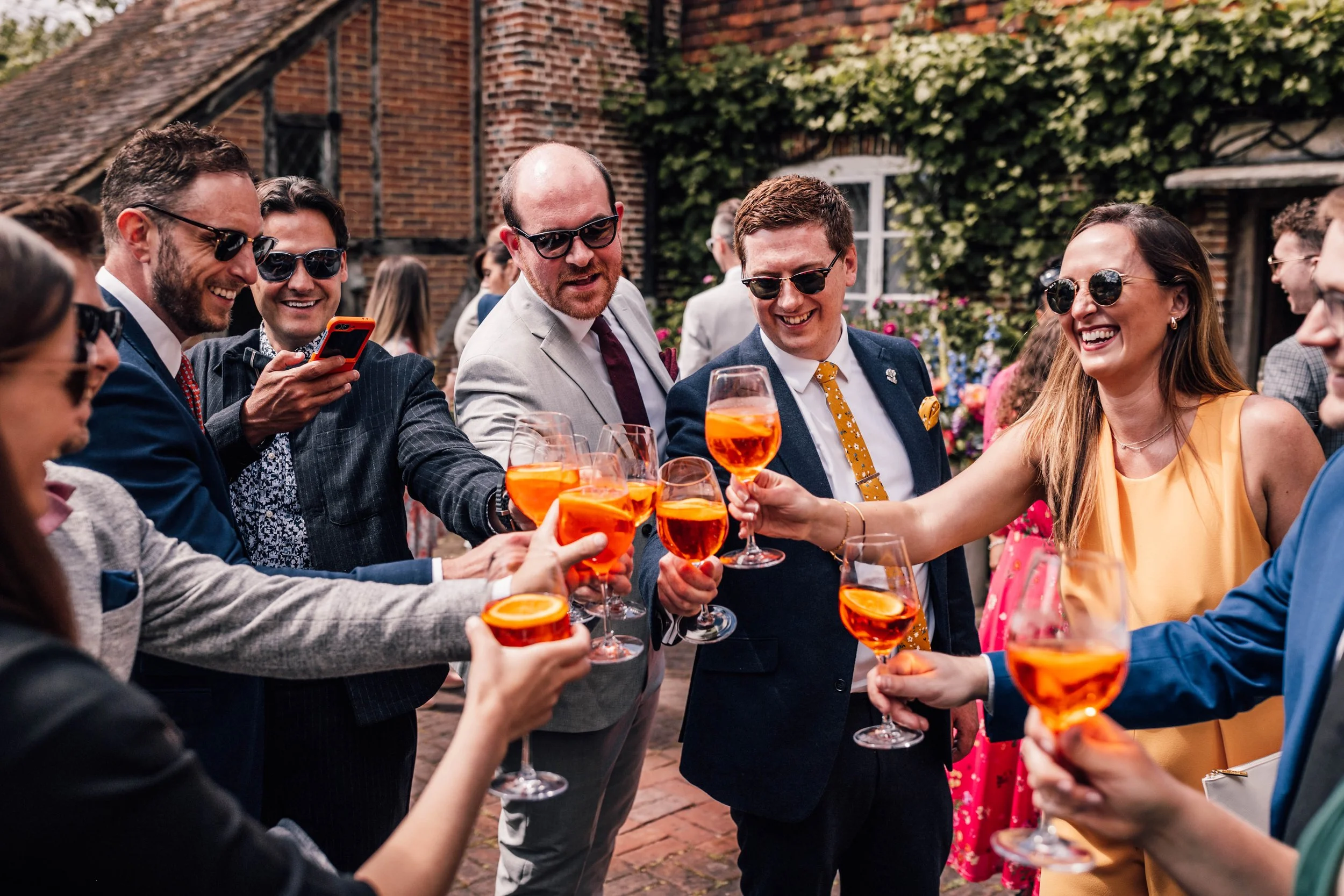 Group of friends at an outdoor celebration clinking glasses with orange-colored drinks, smiling and having a good time.