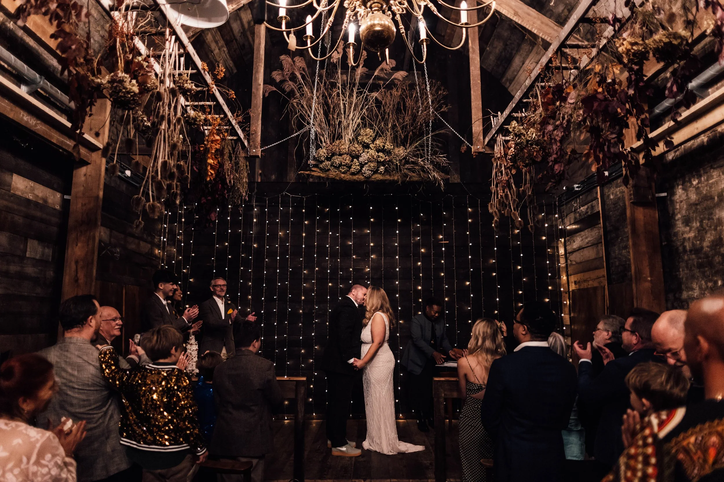A wedding ceremony in a rustic indoor venue with wooden walls and fairy lights, featuring a couple holding hands and kissing at the altar, surrounded by guests clapping and smiling.