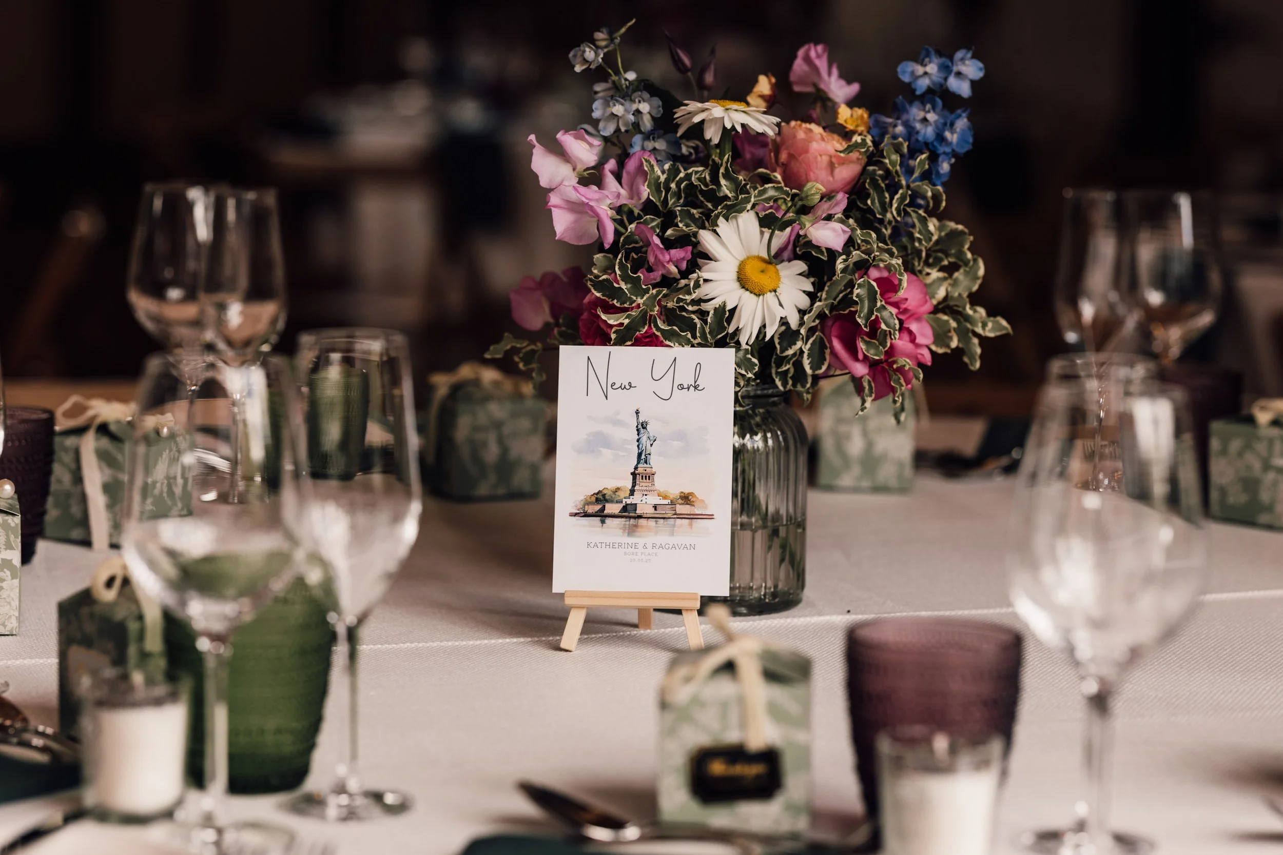Table centerpiece with large flower arrangement of pink, white, and blue flowers, including daisies and roses, in a glass vase, surrounded by glassware, small gift boxes, and candles at a wedding reception.