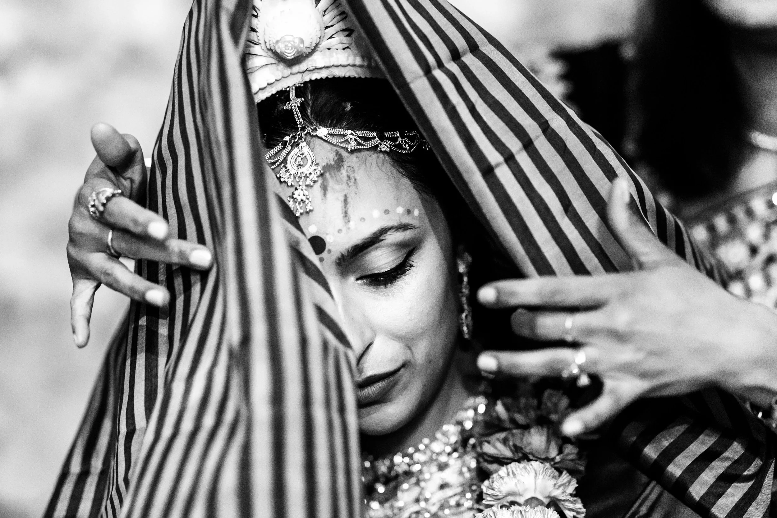 A woman dressed in traditional Indian attire, wearing ornate jewelry and a decorative bindi on her forehead, with her eyes closed and her arms extended around a striped cloth, possibly during a cultural or religious ceremony.