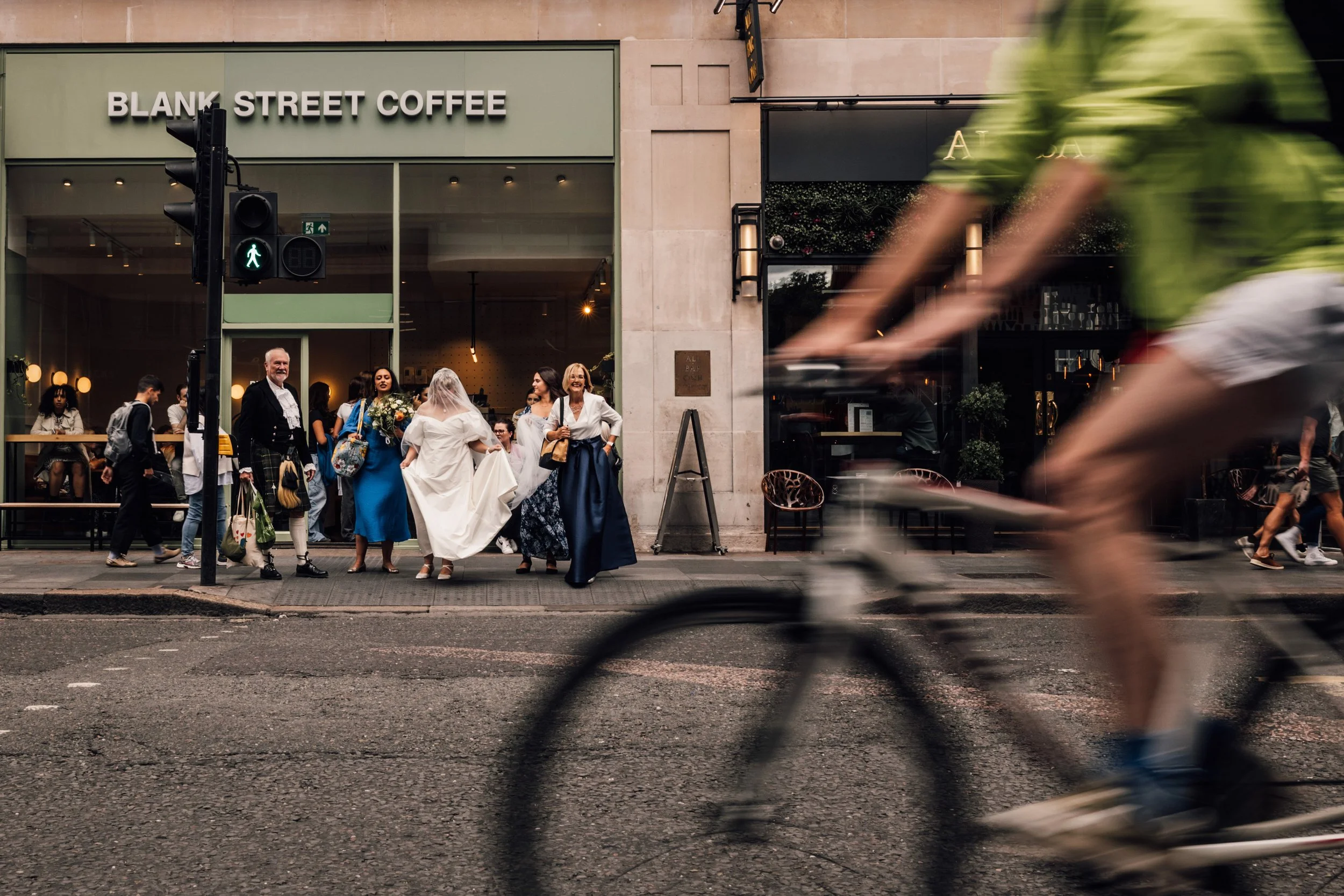 People crossing the street outside a coffee shop called 'Blank Street Coffee.' Some pedestrians are waiting to cross, while a cyclist is riding by in the foreground.