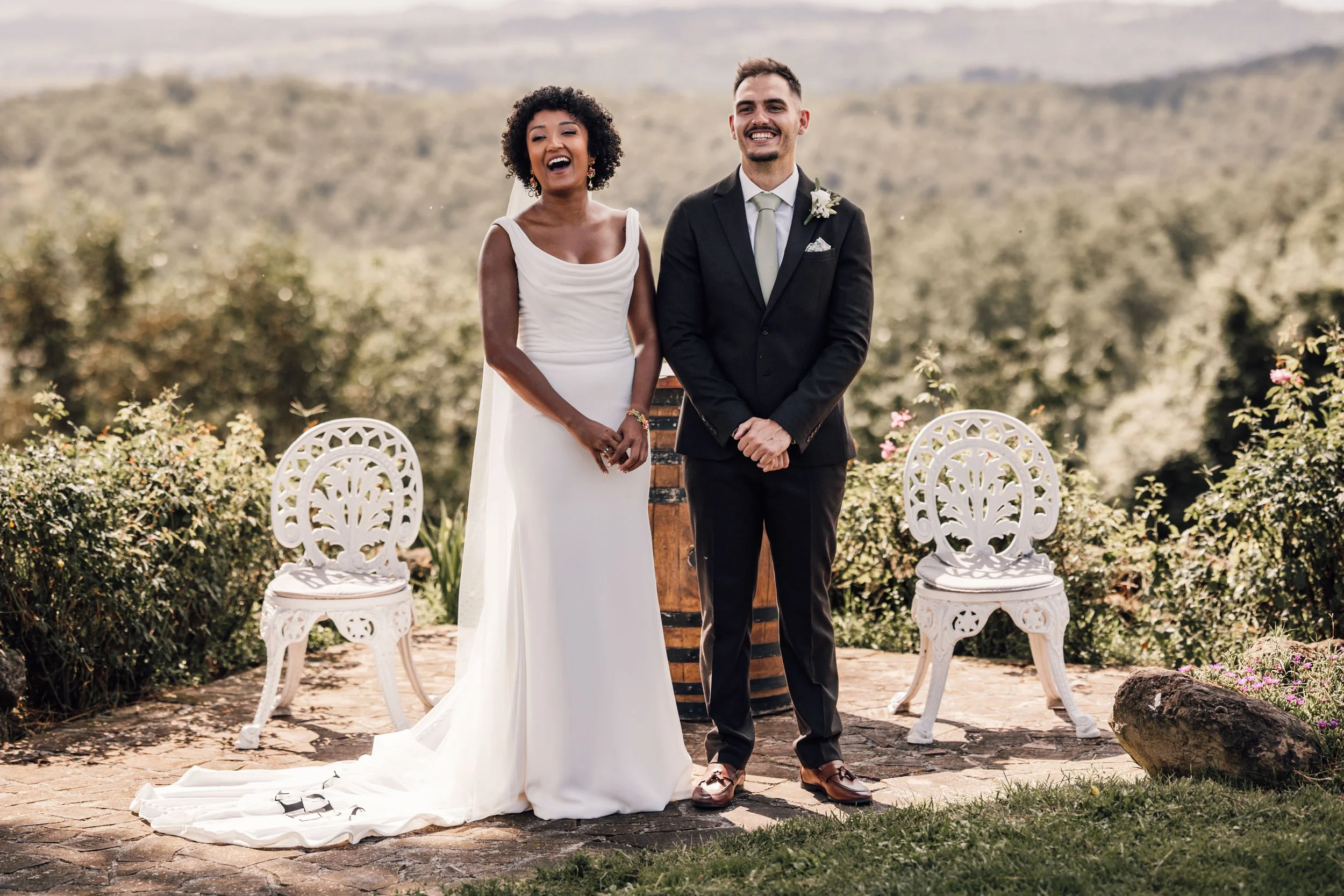 A newlywed couple standing outdoors during their wedding ceremony, smiling, with two white decorative chairs behind them and a lush green landscape in the background.