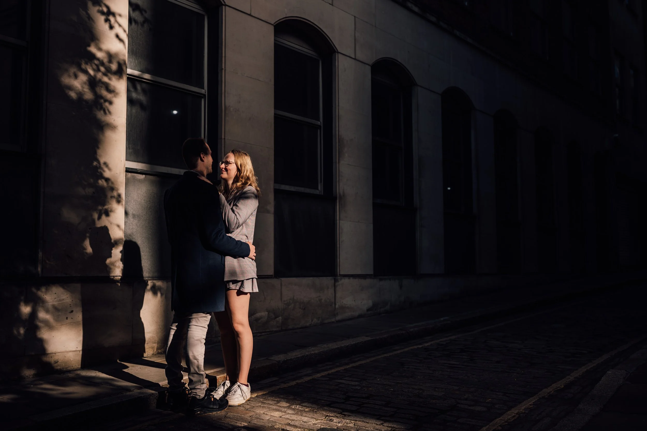 A romantic couple standing on a cobblestone street at night, facing each other and smiling, with a building in the background and shadows cast by nearby trees.