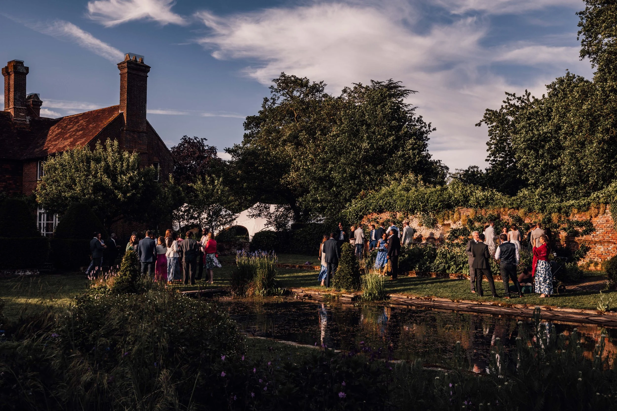 A group of people dressed in formal attire gathering in a garden near a pond, with trees, a brick wall, and a house with brick chimneys in the background under a partly cloudy sky.
