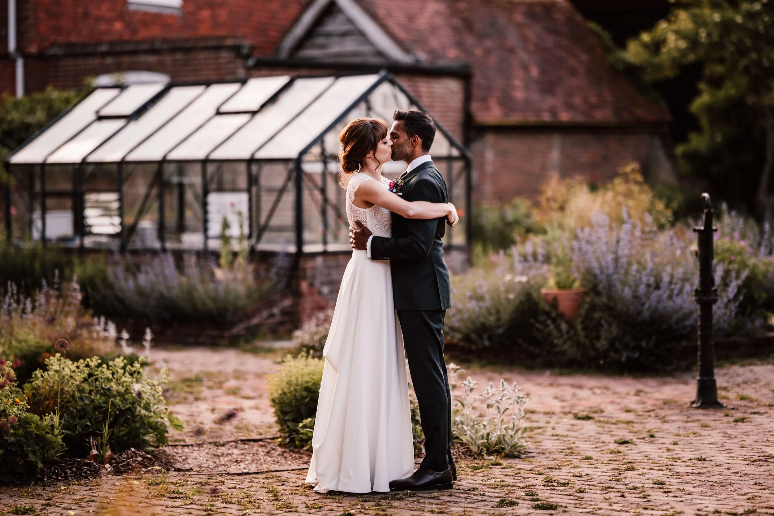 A bride and groom sharing a kiss outdoors in a garden with flowers and a greenhouse in the background.