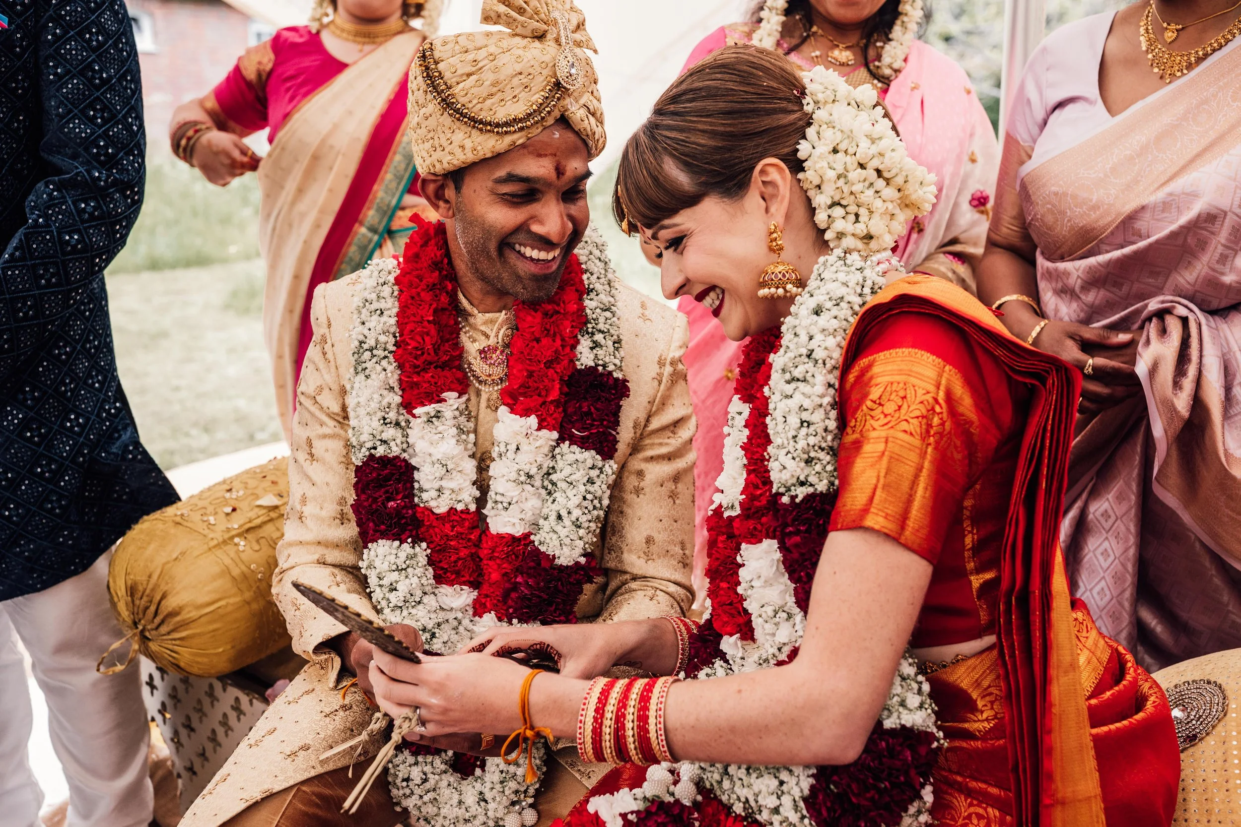 A couple in traditional Indian wedding attire sitting together, smiling and happy, surrounded by women in colorful sarees and jewelry, with garlands of flowers around their necks.