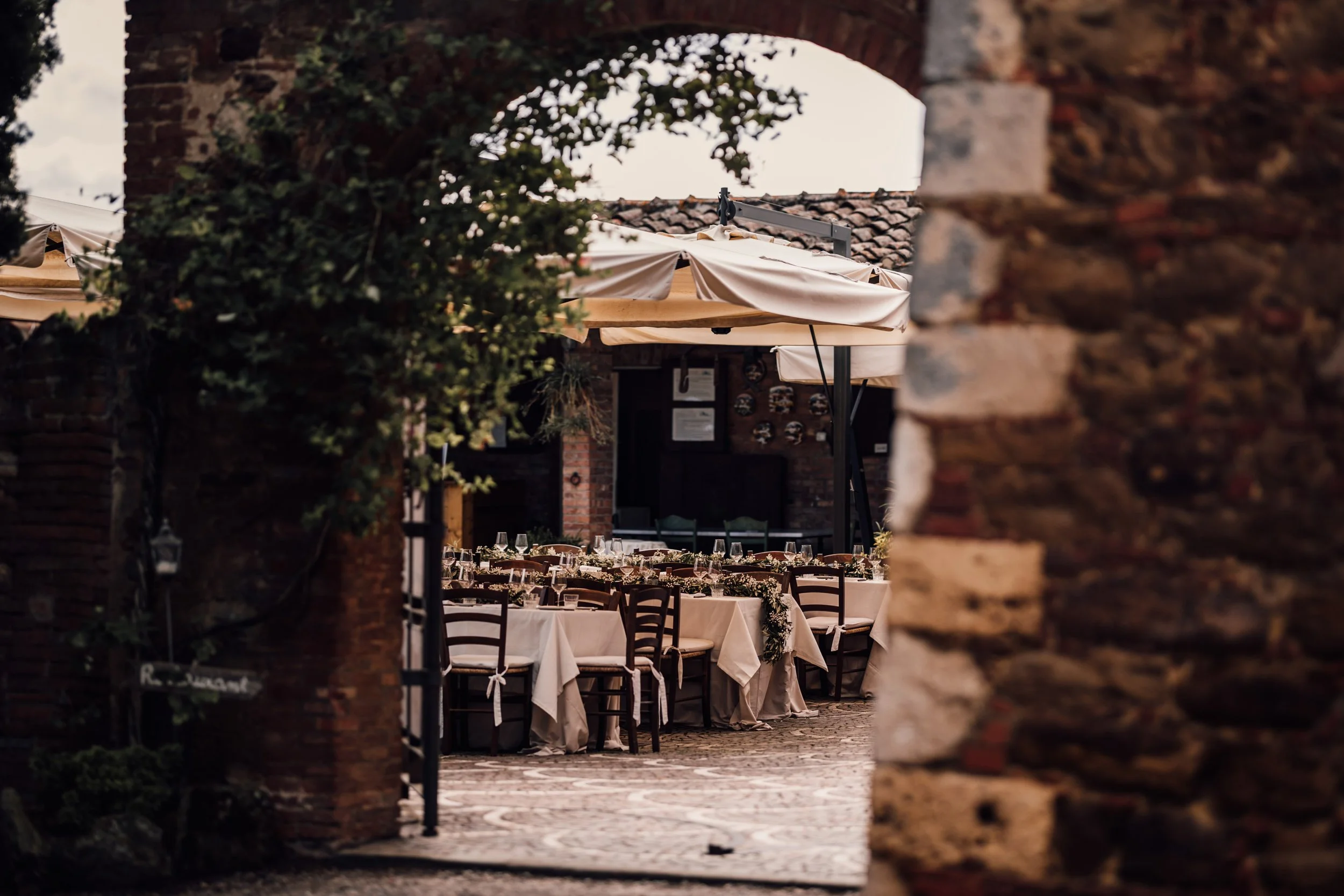 Elegant outdoor dining setup with long tables, wine glasses, and tableware, surrounded by brick walls and large beige umbrellas, creating a cozy atmosphere.