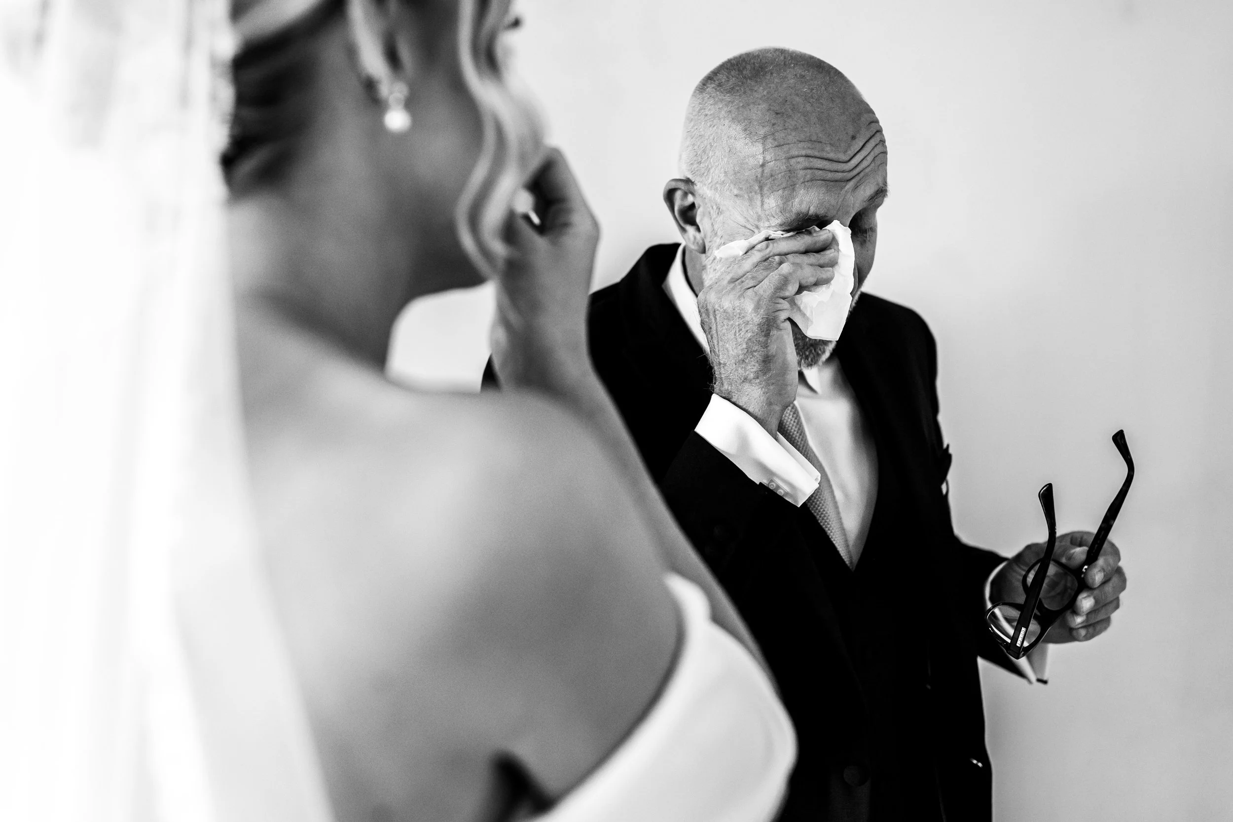 An elderly man in a suit and tie, wiping tears with a tissue, holding glasses, next to a woman in a wedding dress, on a neutral background.