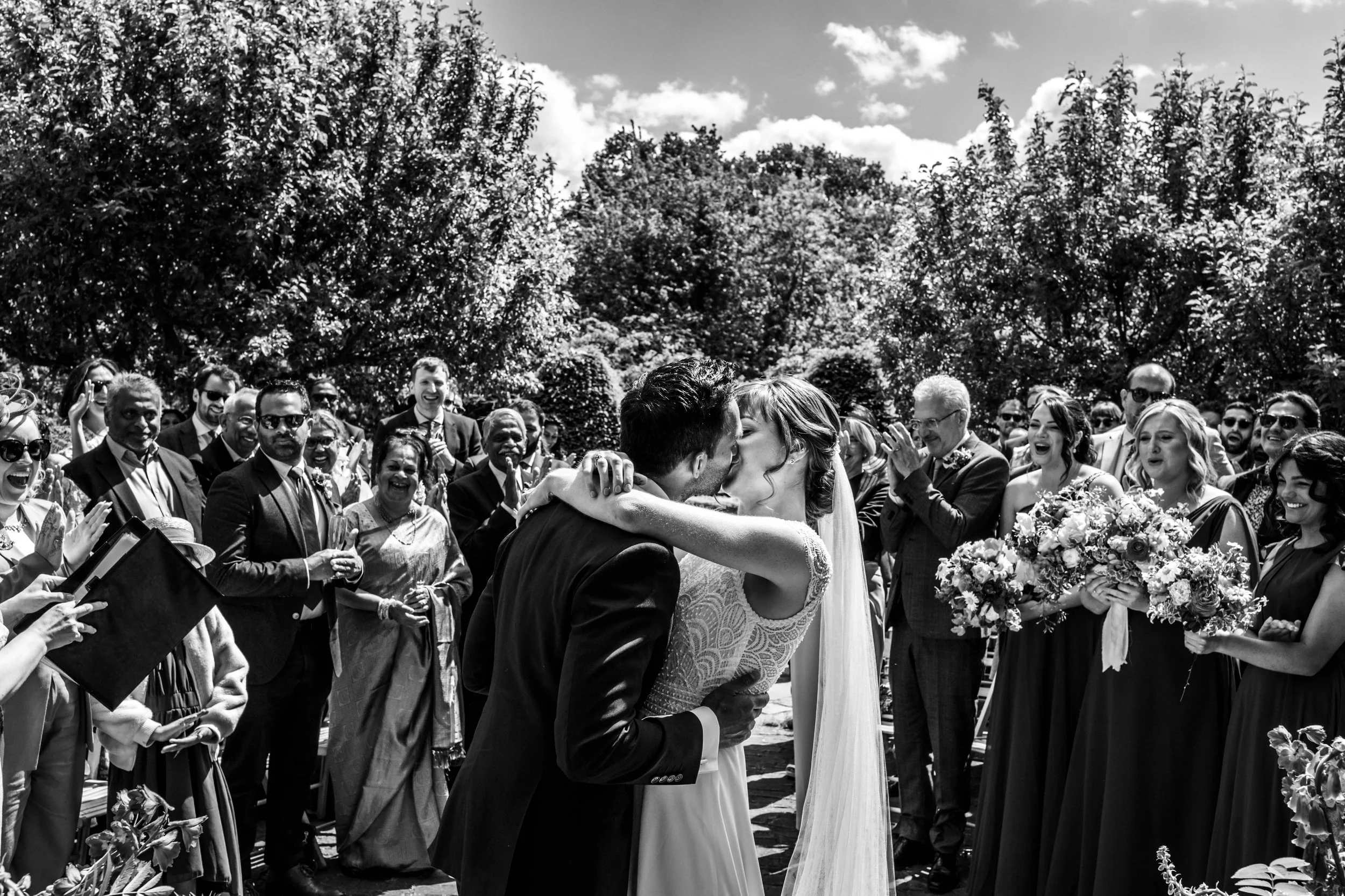 Black and white photo of a wedding ceremony outdoors with a bride and groom kissing while friends and family look on happily.