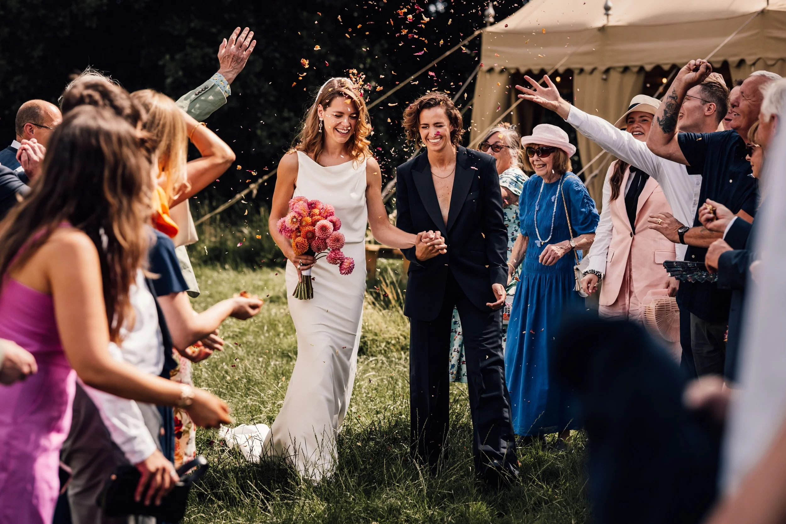 Two women, possibly newlyweds, walking hand in hand through a crowd of people celebrating outdoors. One woman is in a white wedding dress holding a pink bouquet, and the other is in a dark blazer and pants. Guests are throwing confetti and smiling.