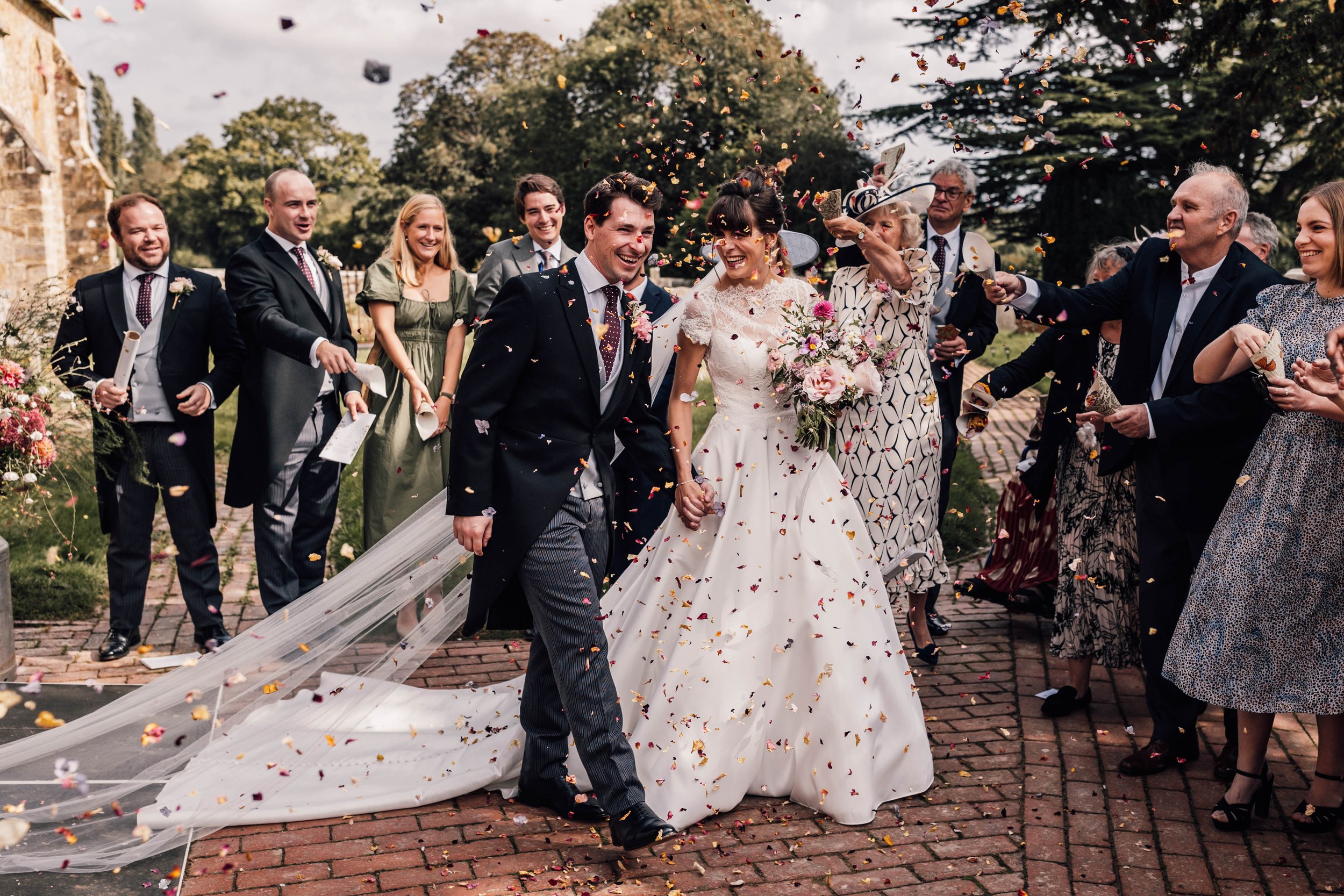 A newlywed couple smiling and holding hands while being showered with confetti by friends and family during their outdoor wedding celebration.