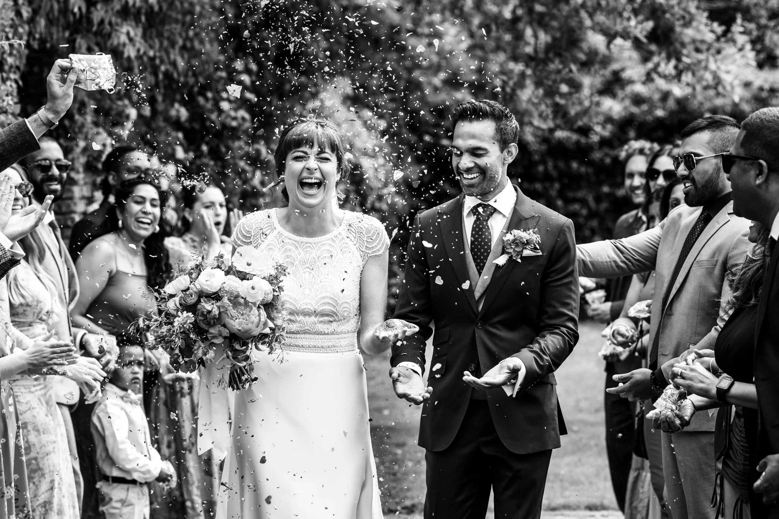 A black and white photo of a wedding celebration outdoors, with a bride and groom surrounded by guests throwing confetti.