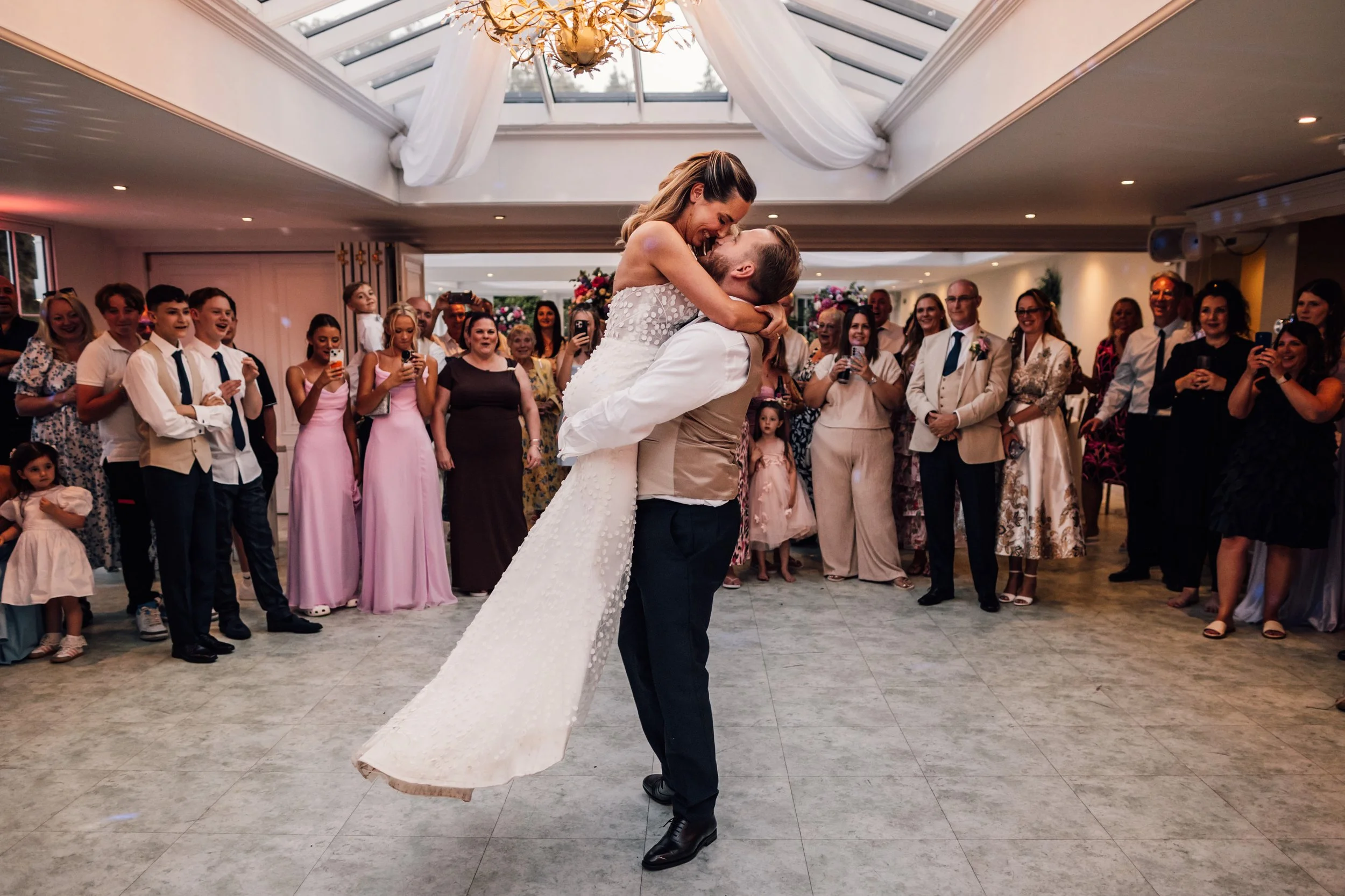 Bride being lifted by groom during wedding dance in reception hall filled with guests taking photos and videos