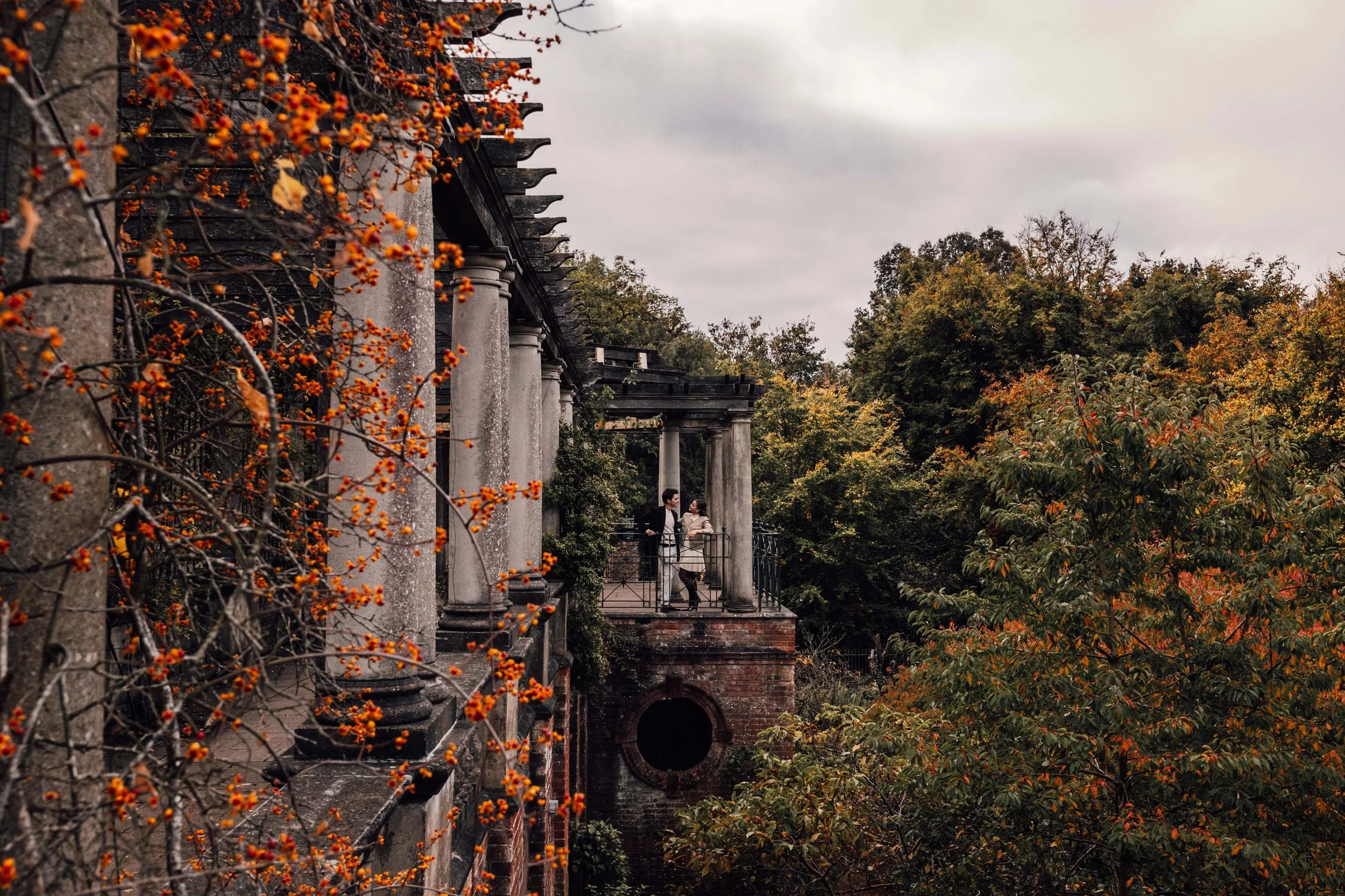 A couple standing on a balcony with grey pillars surrounded by autumn trees with orange and green leaves.