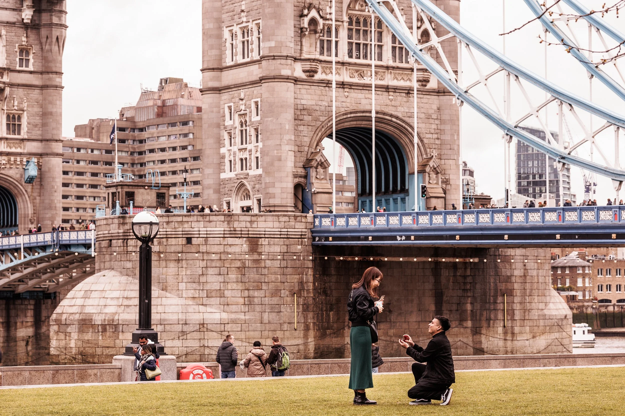 A man proposing to a woman near Tower Bridge, London, with people in the background.